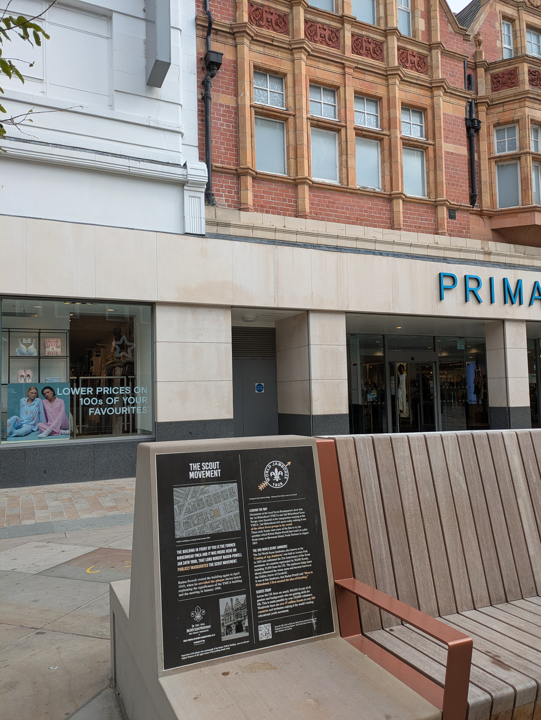A public bench with an informational plaque about The Scout Movement is positioned in front of a Primark store and adjacent buildings.