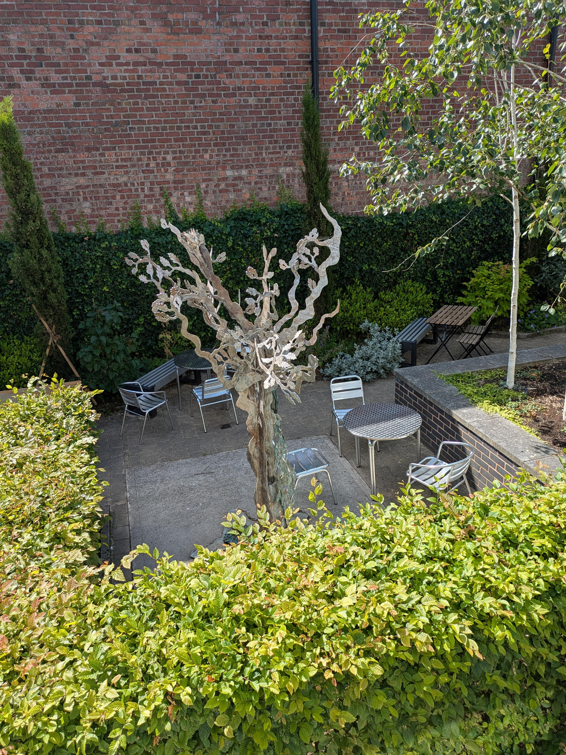 A courtyard features a metal sculpture resembling a tree surrounded by metal chairs and tables, with greenery and a brick wall in the background.