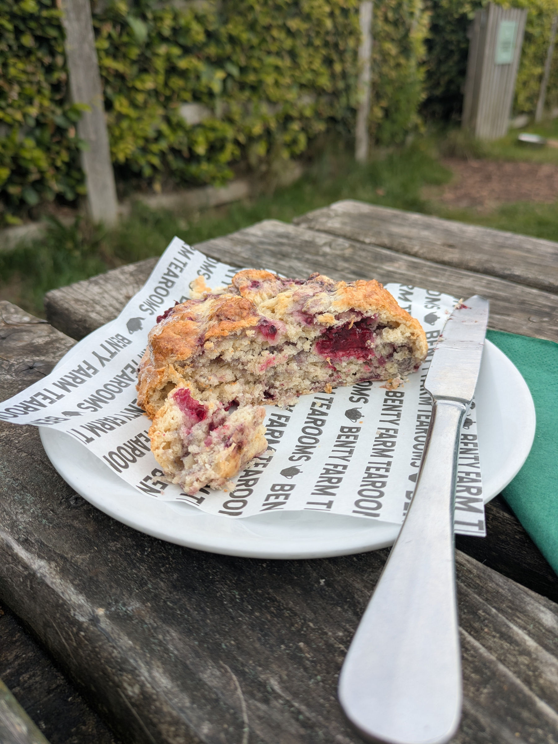 A partially eaten slice of tayberry scone is placed on a white plate with branded paper, accompanied by a butter knife, on a rustic wooden outdoor table.