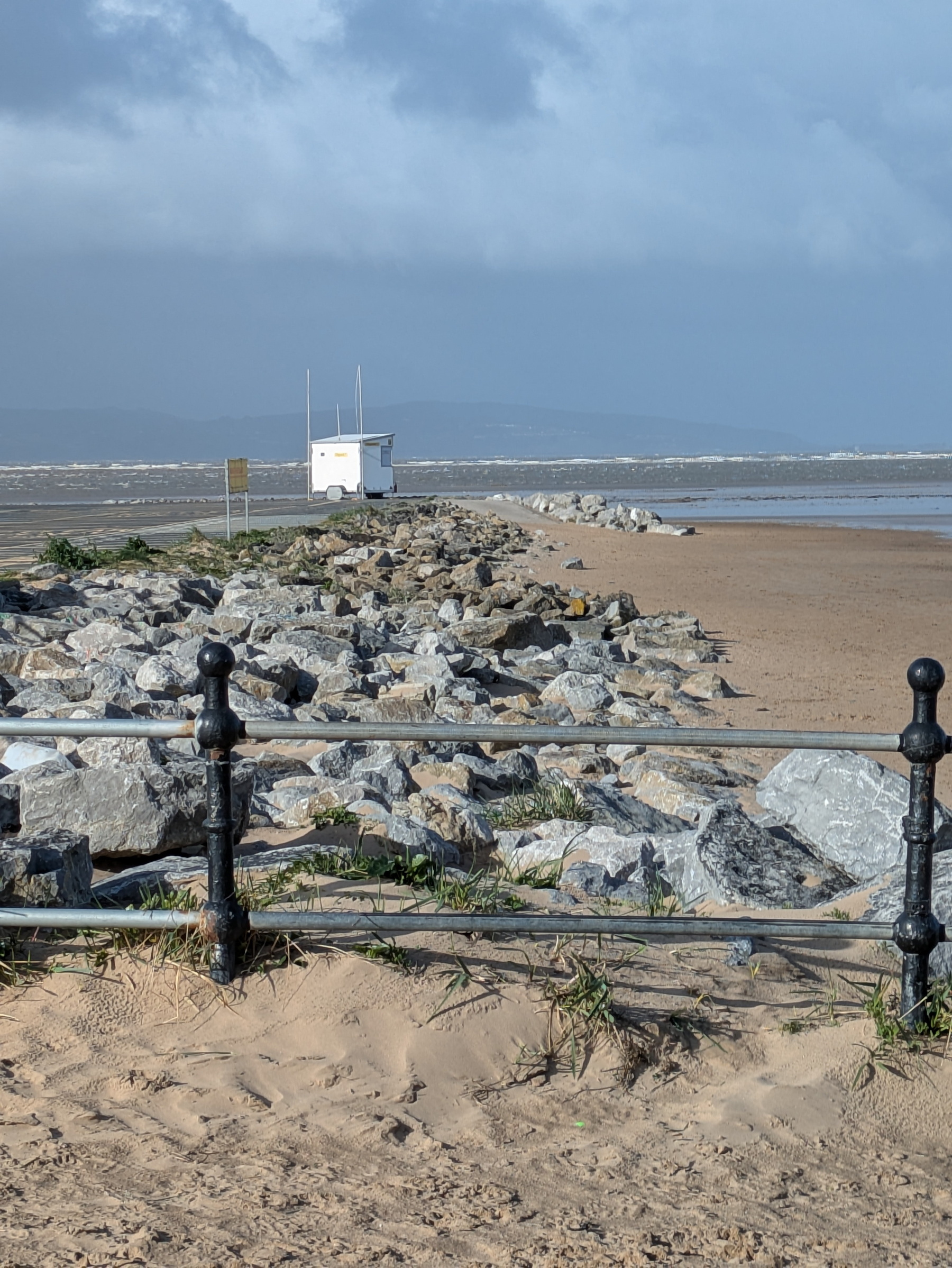 A rocky shoreline with a small white structure in the distance, bordered by a metal railing, is set against a cloudy sky.