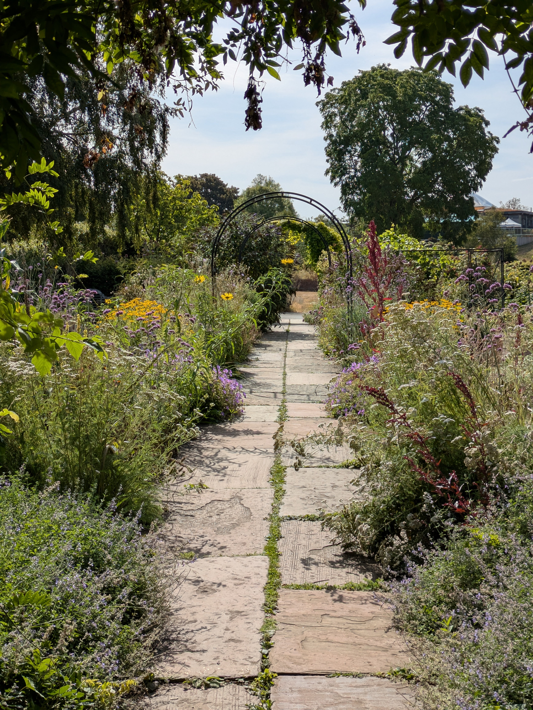 A stone pathway bordered by lush, colorful flowers and greenery leads to a metal archway under a partly cloudy sky.