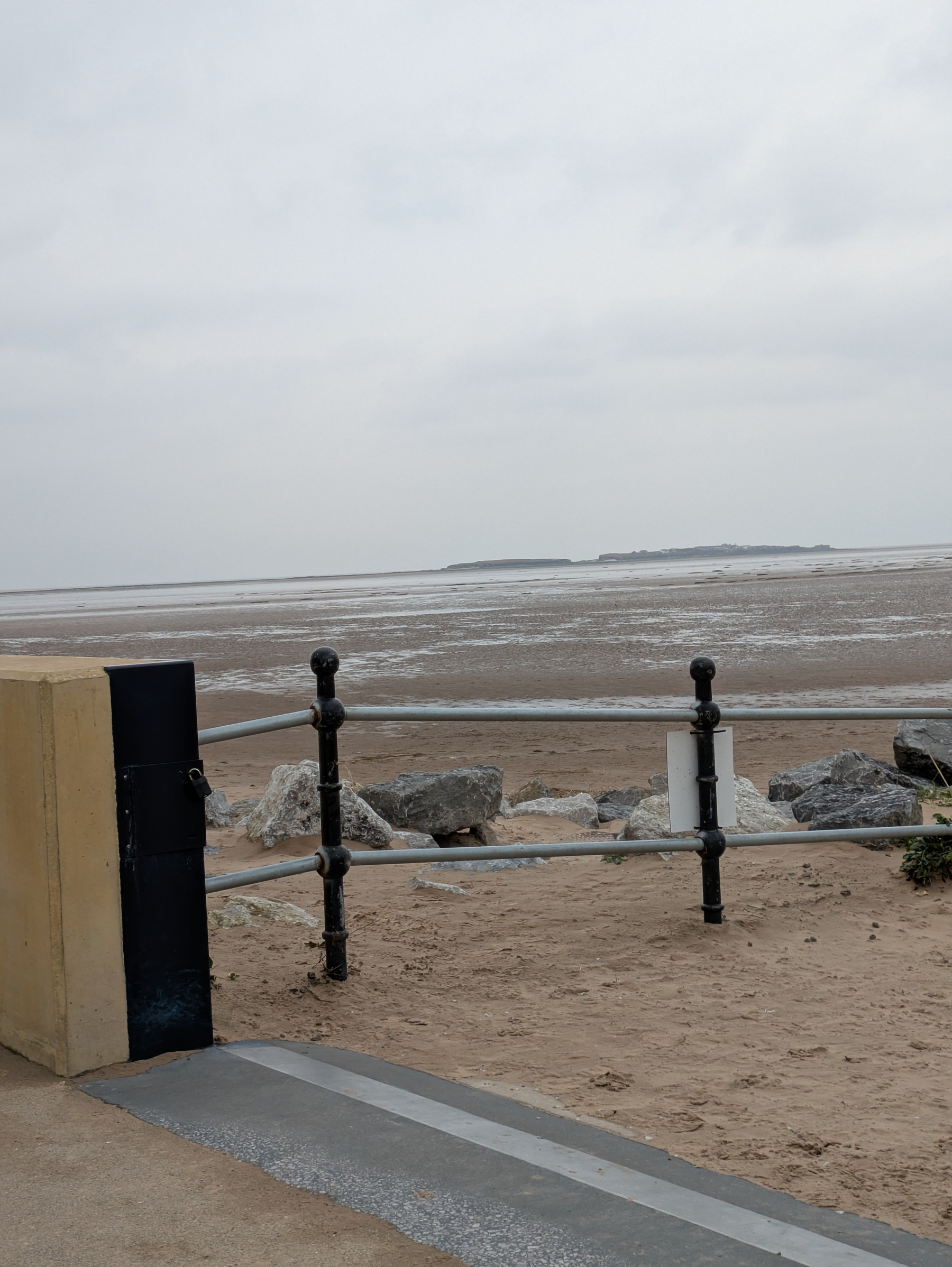 A sandy beach stretches out to the horizon under a cloudy sky, with a fenced walkway and rocky foreground.