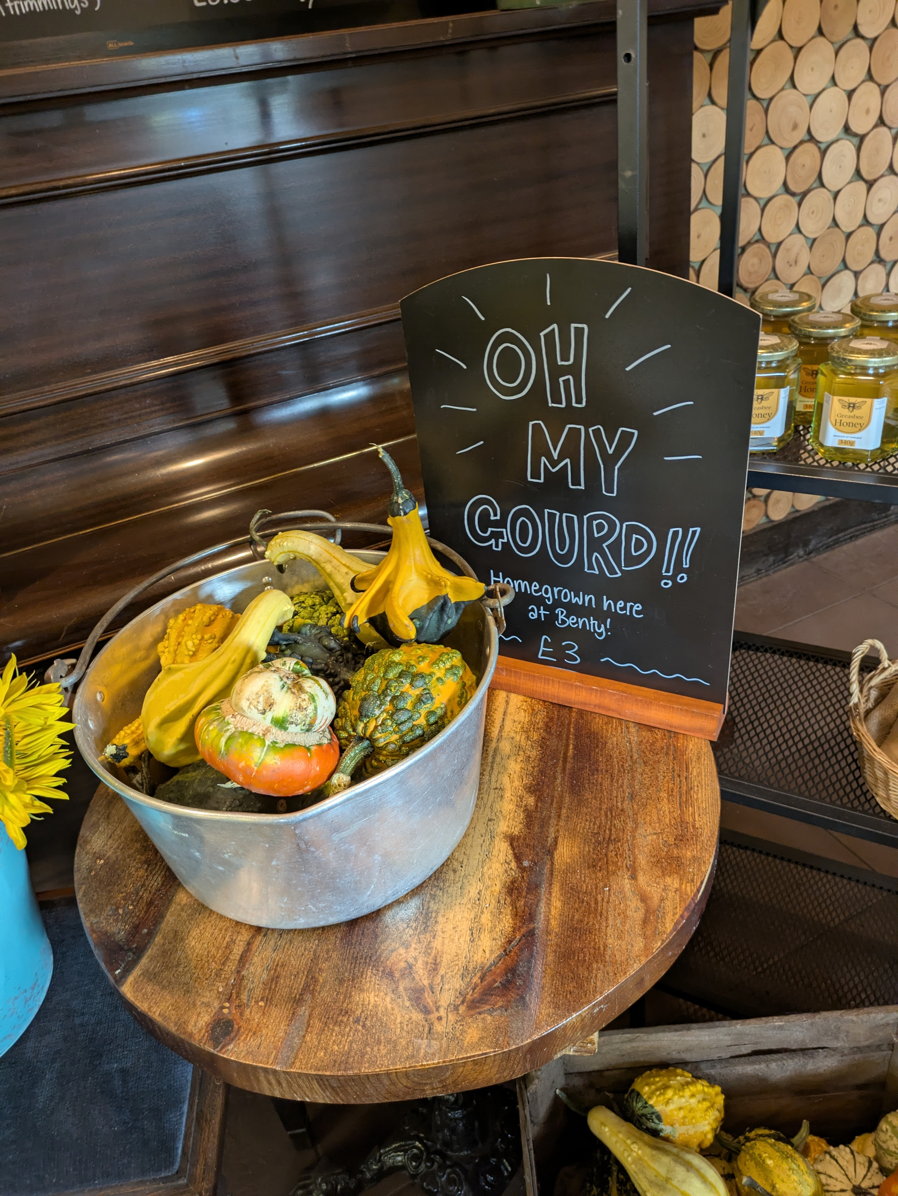 A metal tub filled with various colorful gourds is displayed on a wooden table next to a handwritten sign promoting homegrown gourds for £3.