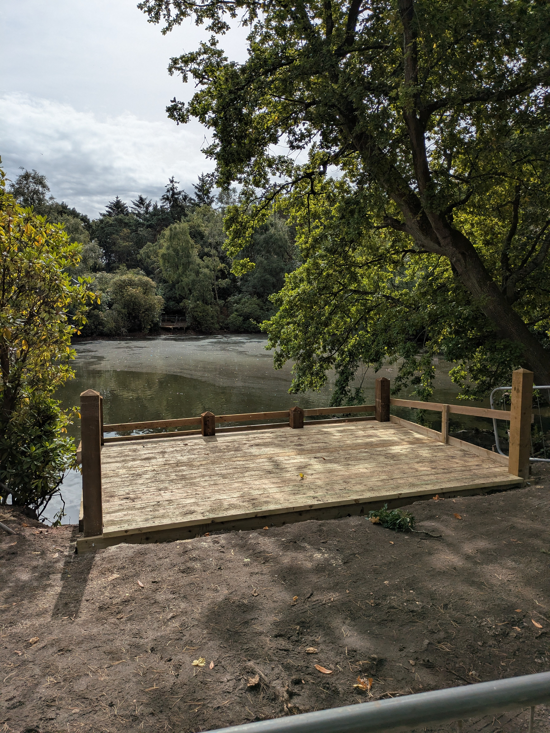 A wooden platform or deck is situated by the edge of a calm, tree-lined body of water on a partly cloudy day.