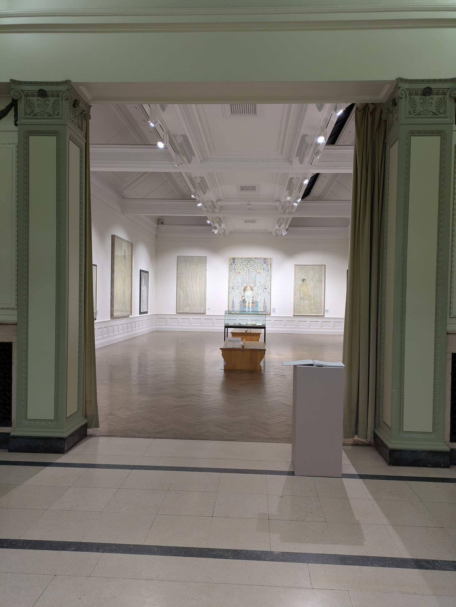 A spacious art gallery room with ornate architectural details features paintings on the walls and display cases in the center.