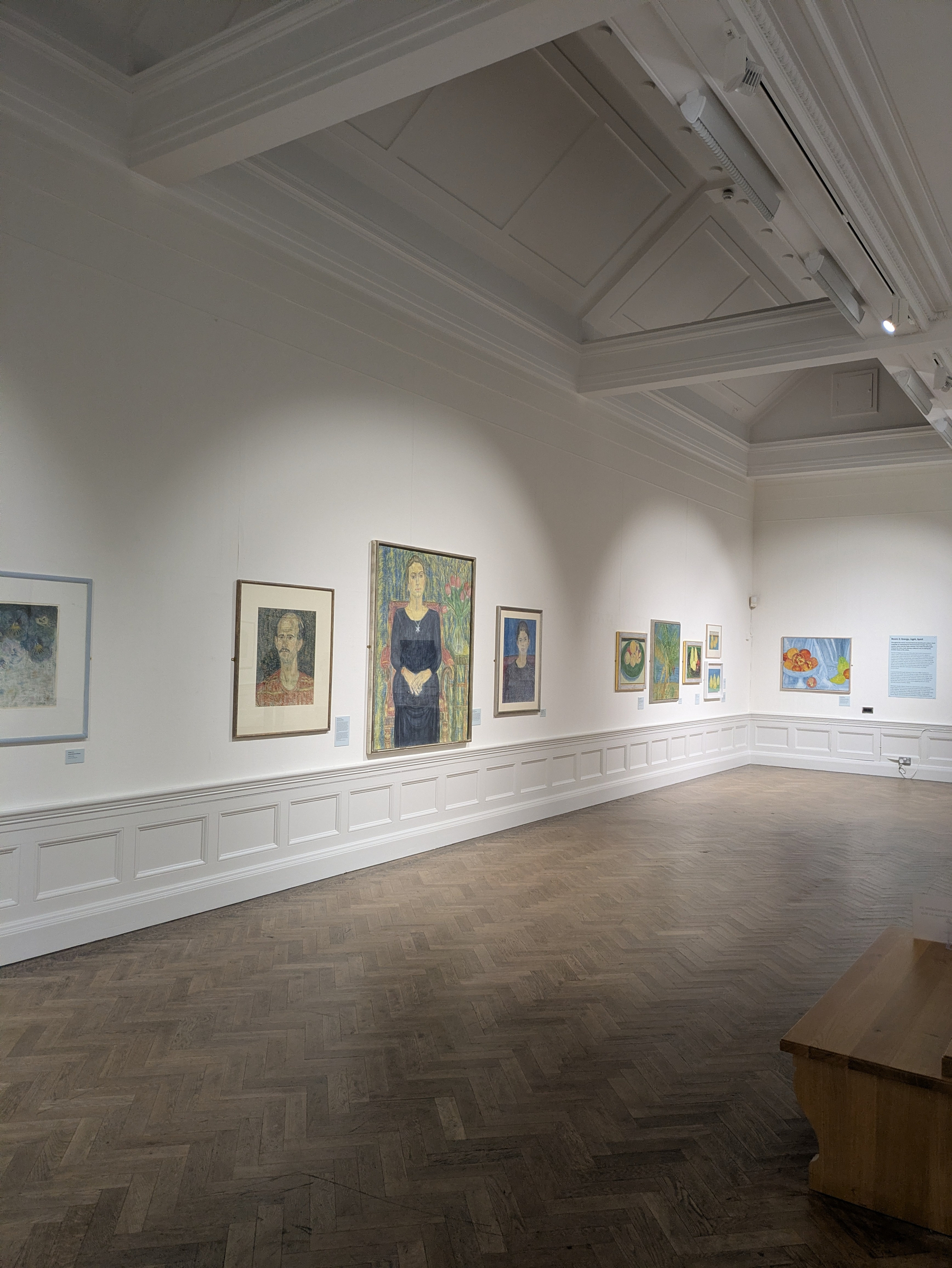 A well-lit art gallery features a collection of framed paintings displayed on white walls with a wooden bench positioned on the right side of the room.