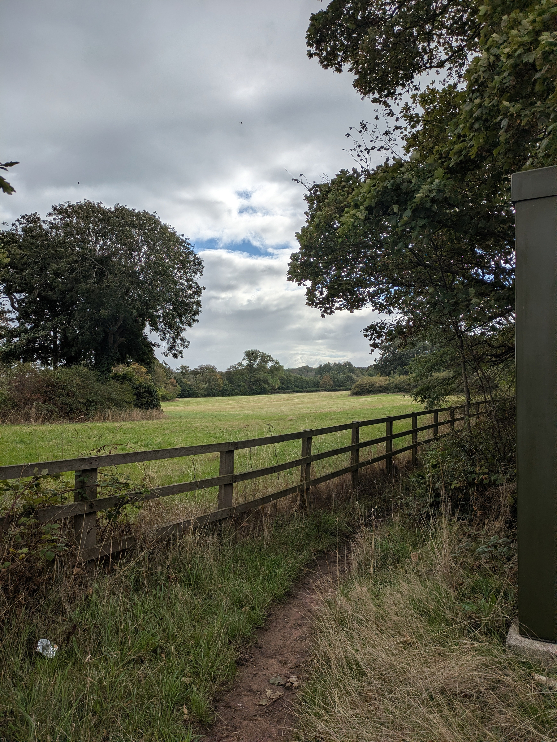 A narrow dirt path runs alongside a wooden fence bordering an open grassy field under a cloudy sky.