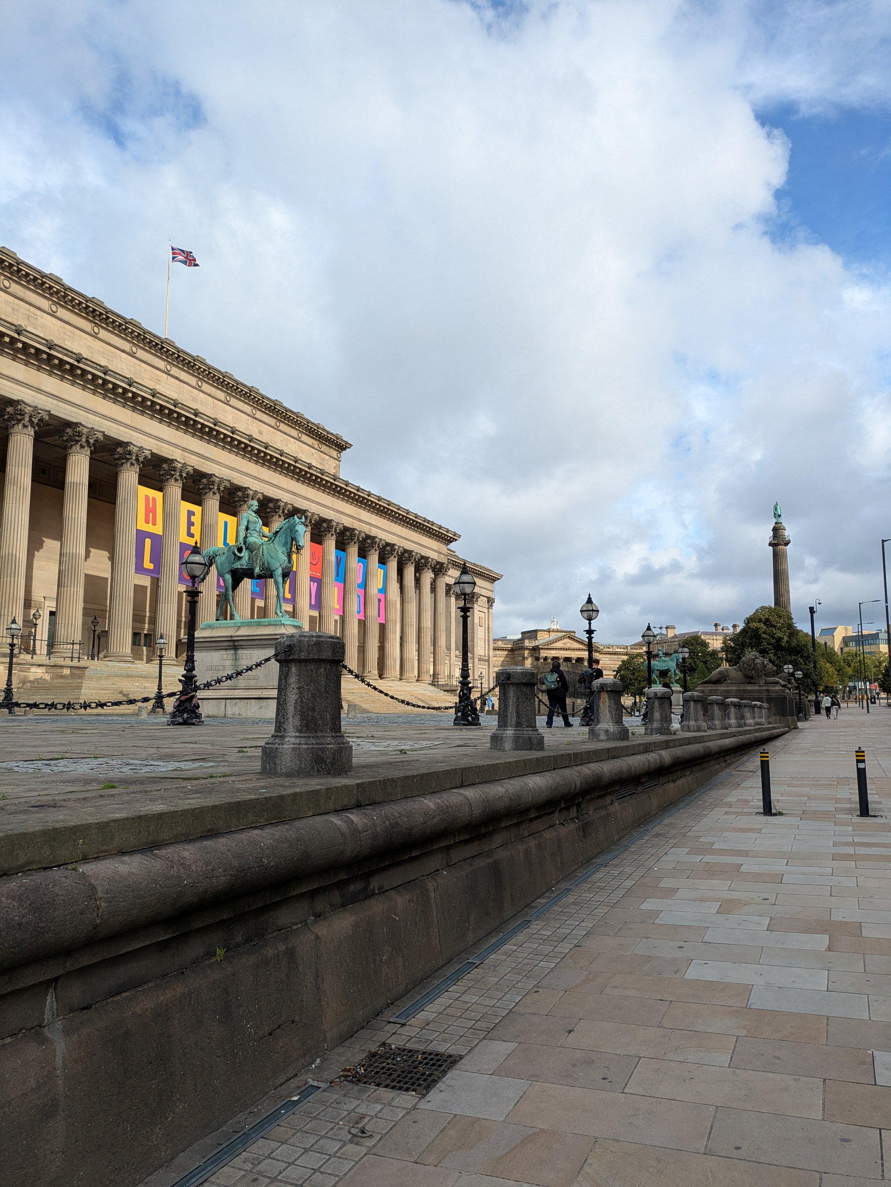 A historic building with large columns, colorful banners, and a statue of a person on horseback is set against a partly cloudy sky and an open paved walkway.