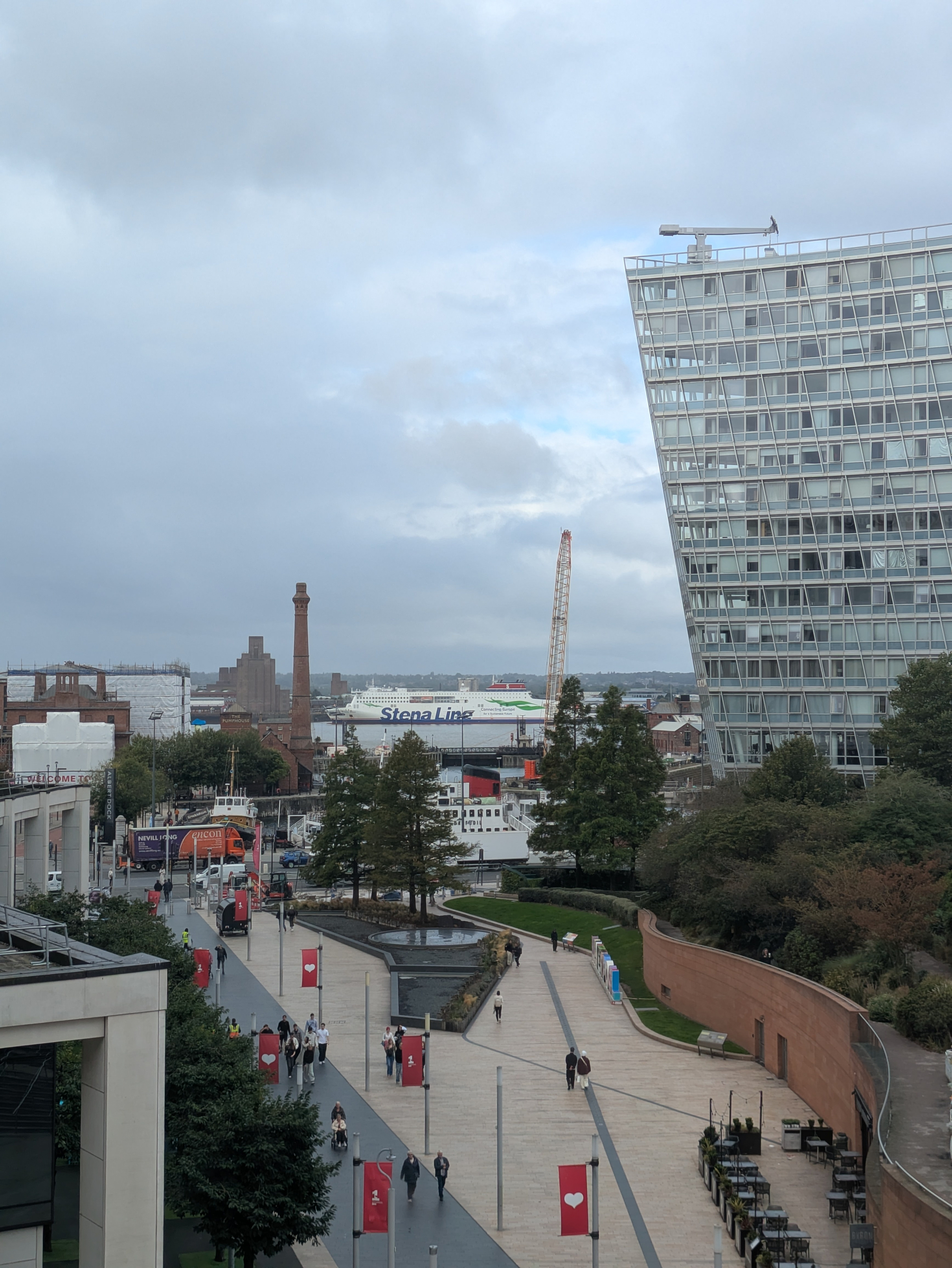 A modern urban scene features pedestrians walking along a paved pathway lined with trees and red banners, with a large glass building and industrial structures visible in the background. Site of the Old Dock. 