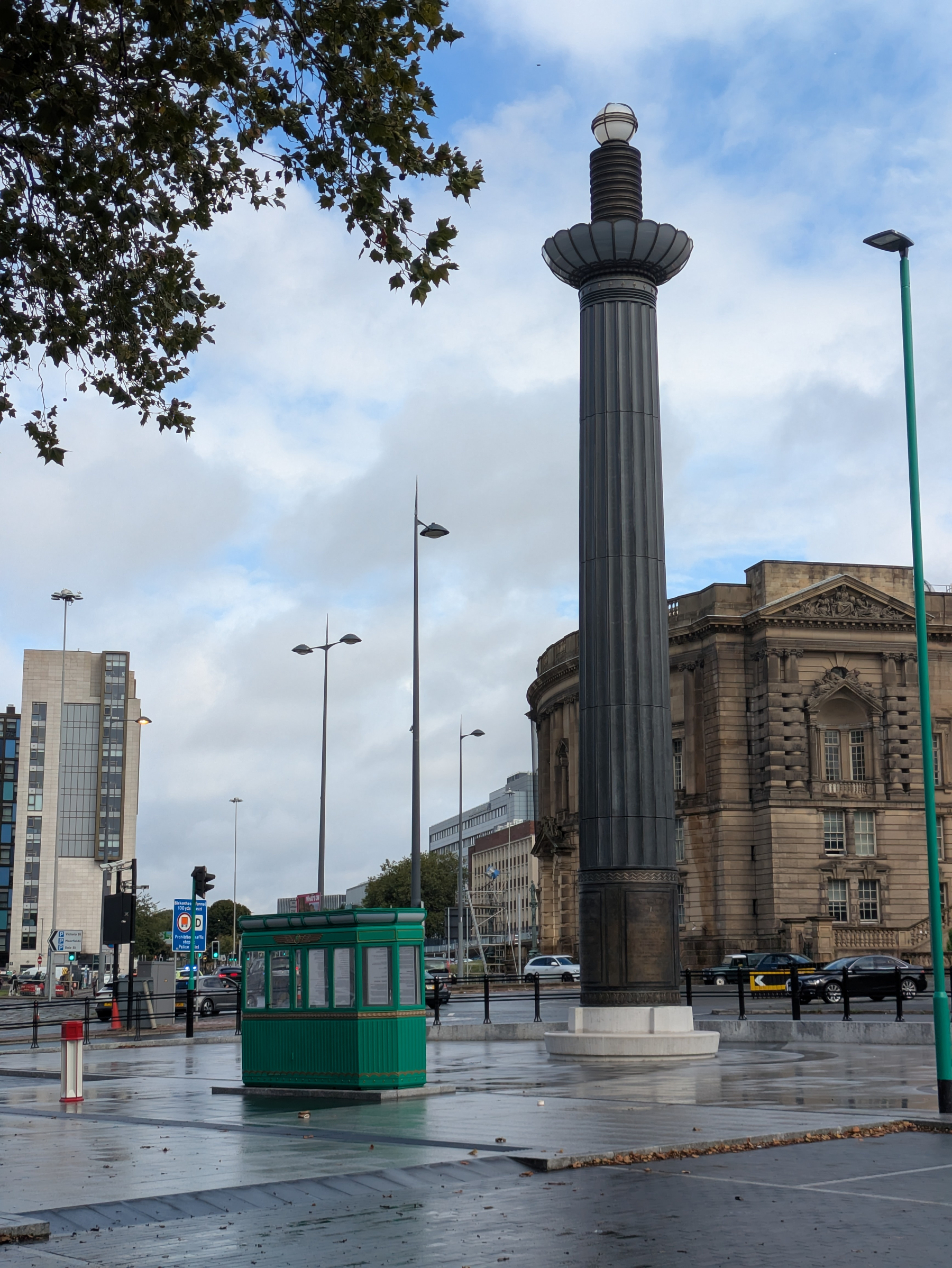 A tall, dark column-like monument stands prominently at the entrance to one of the Mersey tunnels on a wet city street corner with surrounding buildings and streetlights under a cloudy sky.