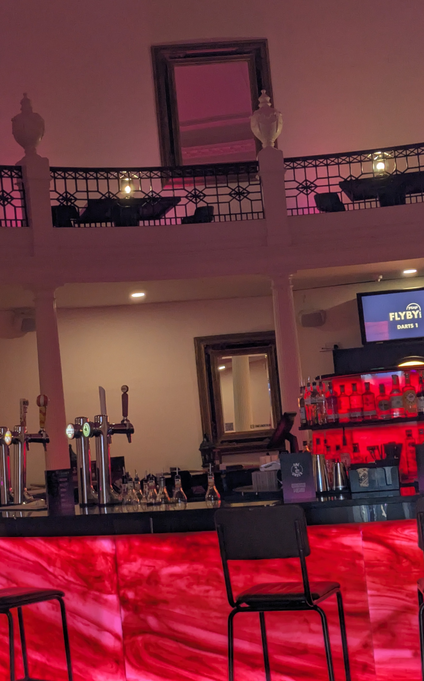 A bar inside the Lyceum with a glowing red countertop, black stools, drink taps, and framed mirrors on the wall is shown in a dimly lit setting.