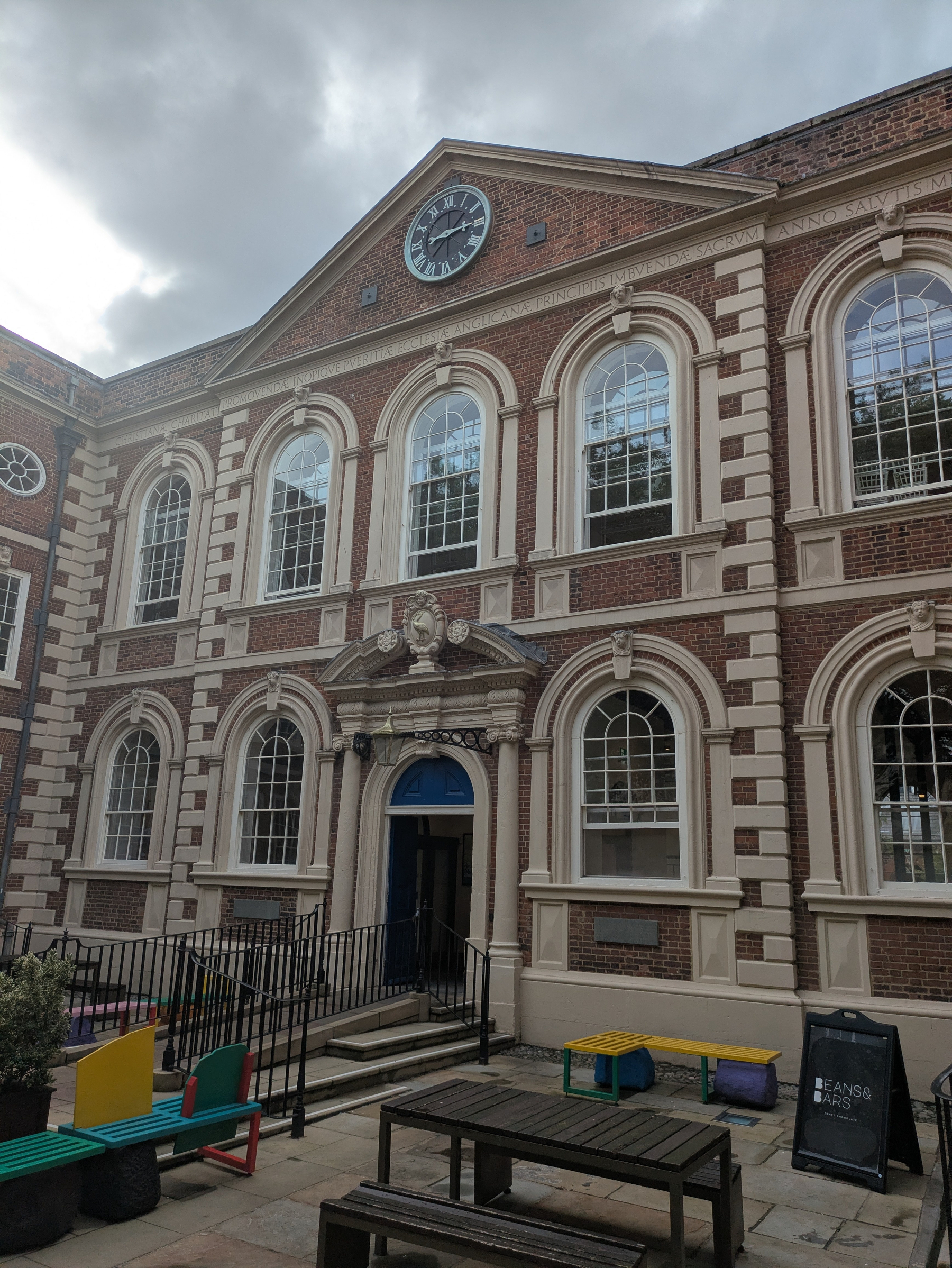 A historic brick building, the Bluecoat, with arched windows and a clock above the entrance overlooks an outdoor seating area with colorful benches and a signboard.