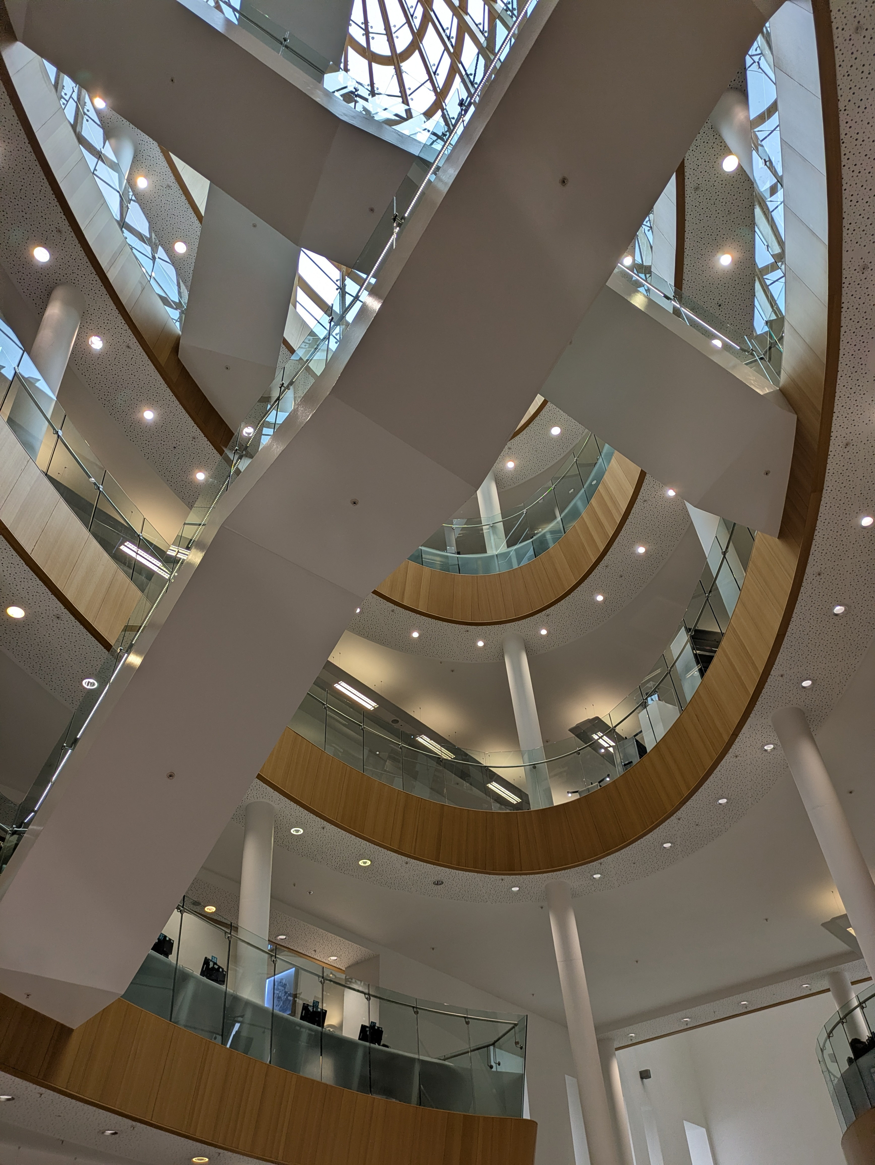 A modern multi-level atrium in the central library with open walkways, glass railings, white pillars, and wooden accents is illuminated by natural light from large skylights.
