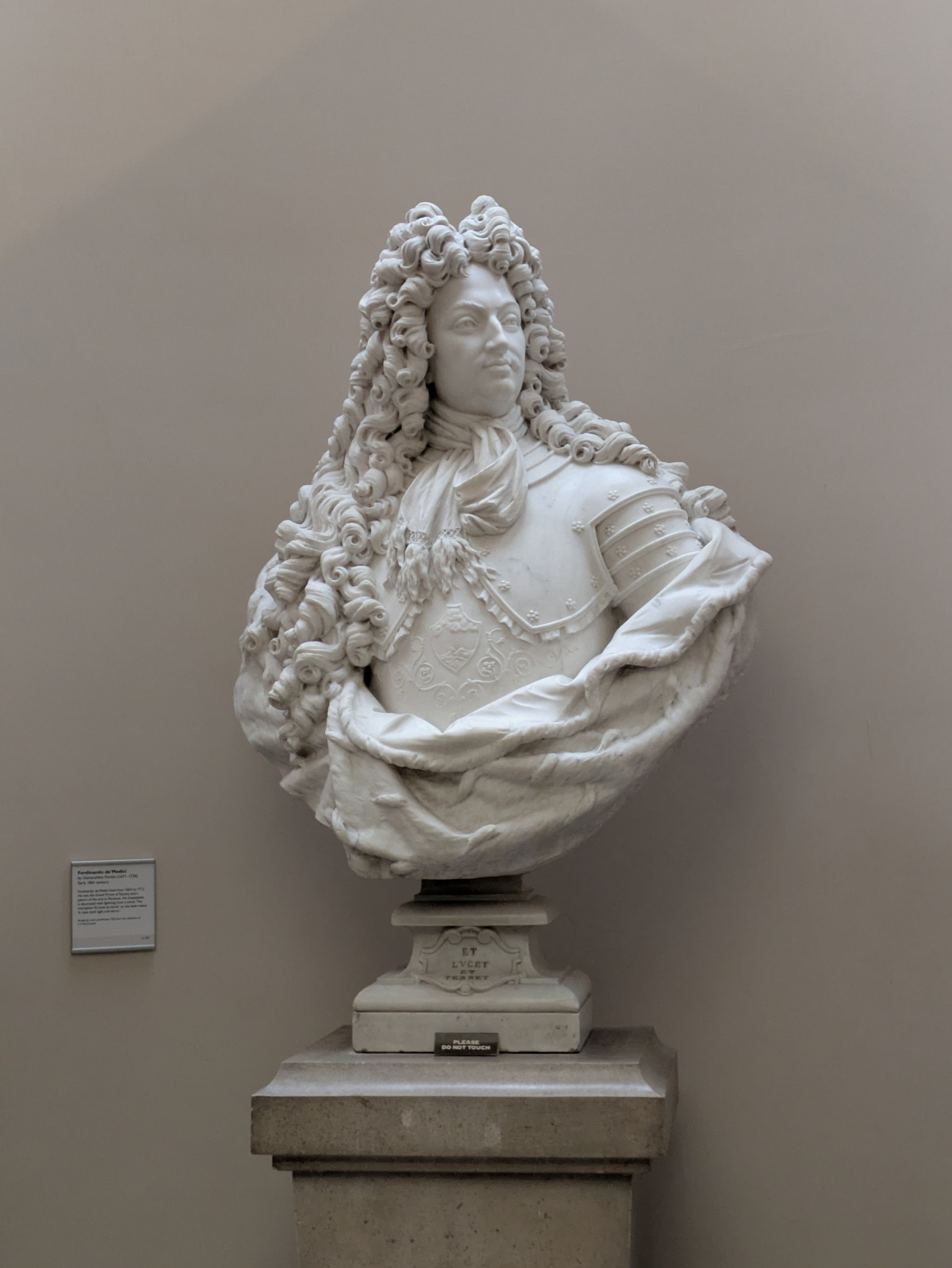 A detailed marble bust of a man with long curly hair, wearing armor and a draped garment, is displayed on a pedestal against a plain background.