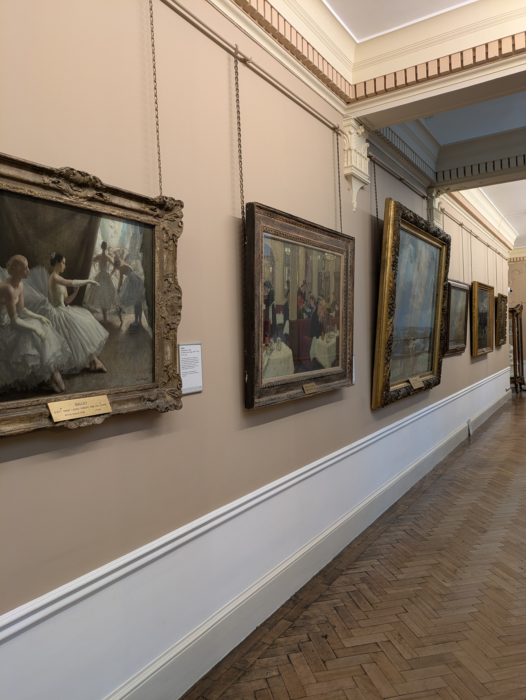 A gallery hallway displays a series of framed paintings hung on beige walls with a wooden herringbone floor beneath.