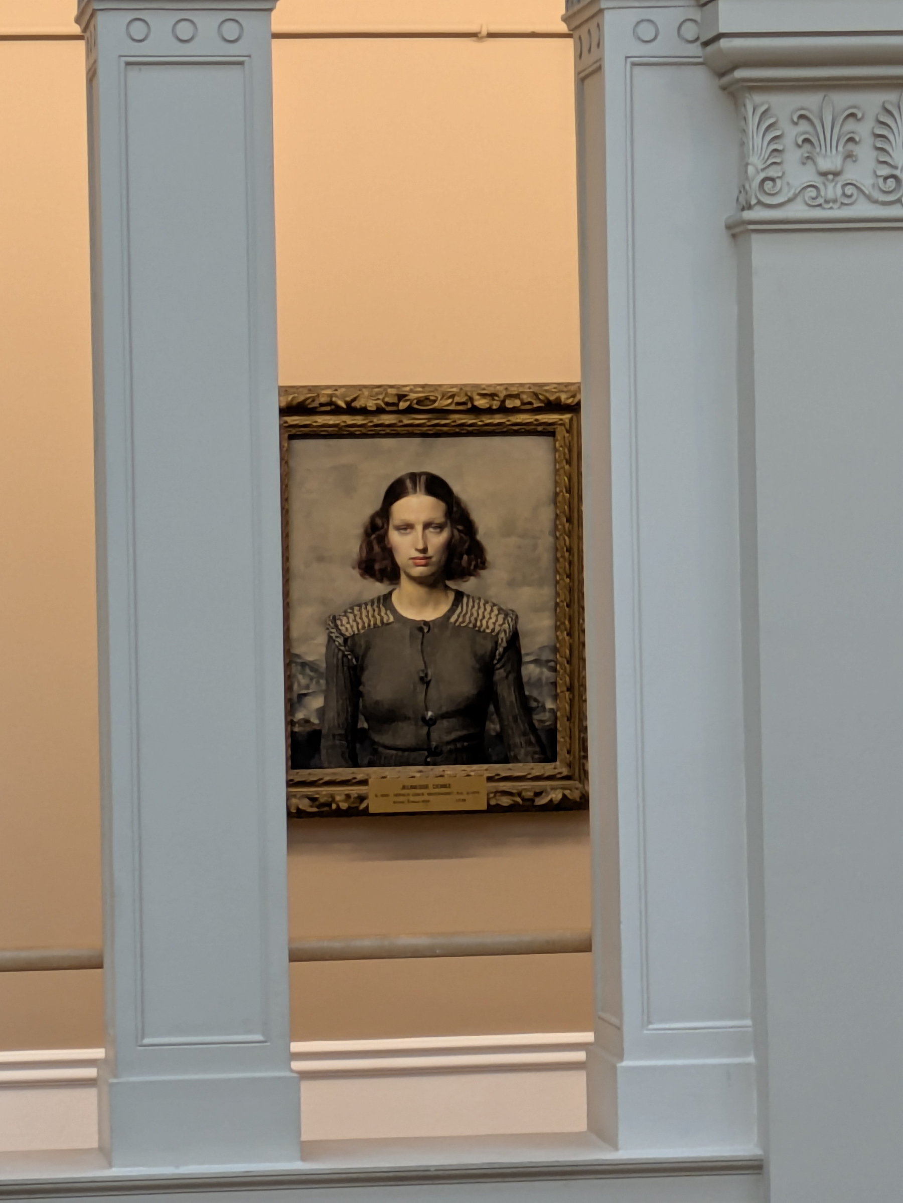 A framed portrait of a woman with dark hair, wearing a buttoned black top, is displayed on a beige wall, partially viewed through a white architectural column.