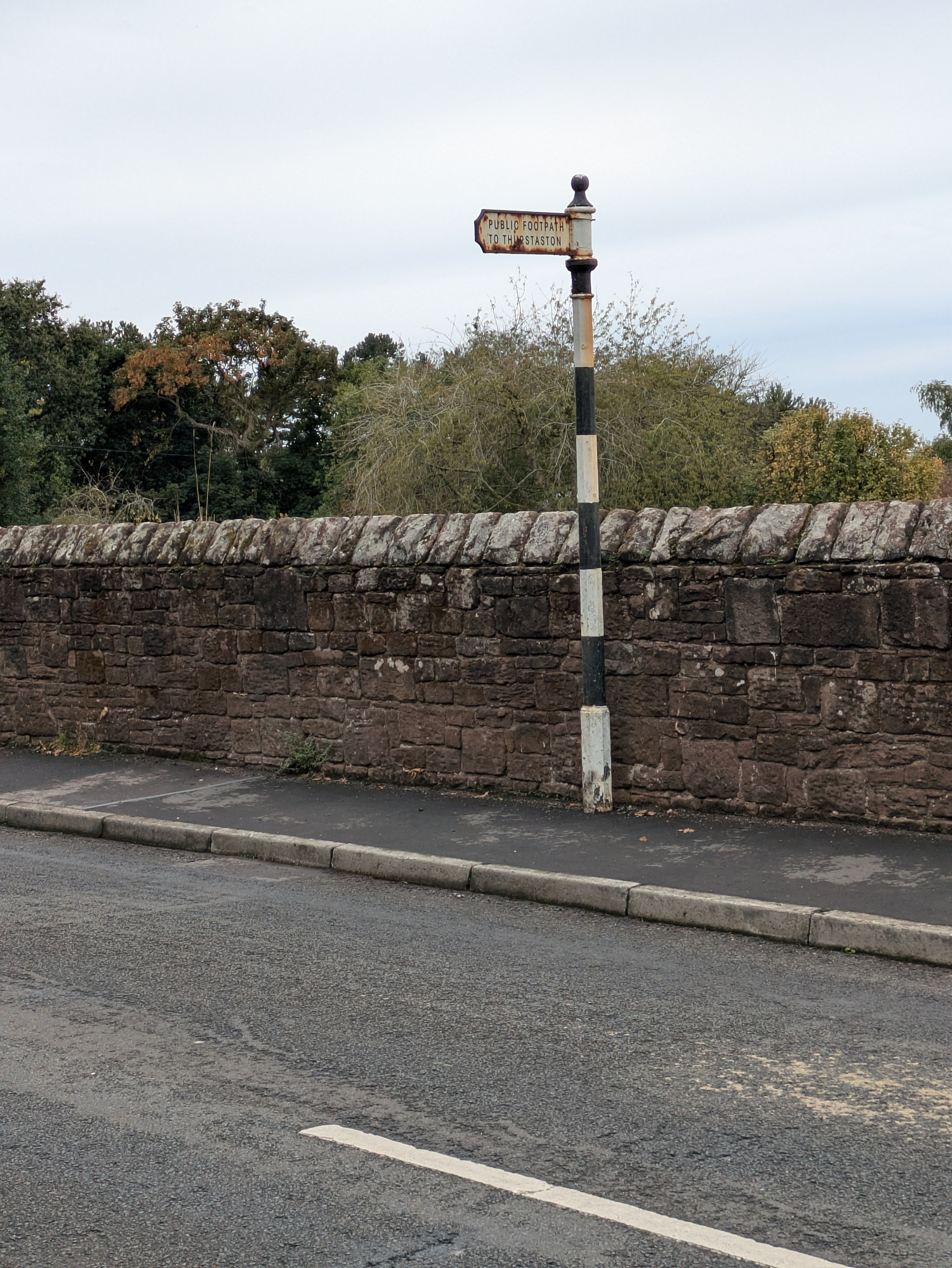 A black and white striped signpost indicating a public footpath stands next to a stone wall along a roadside.
