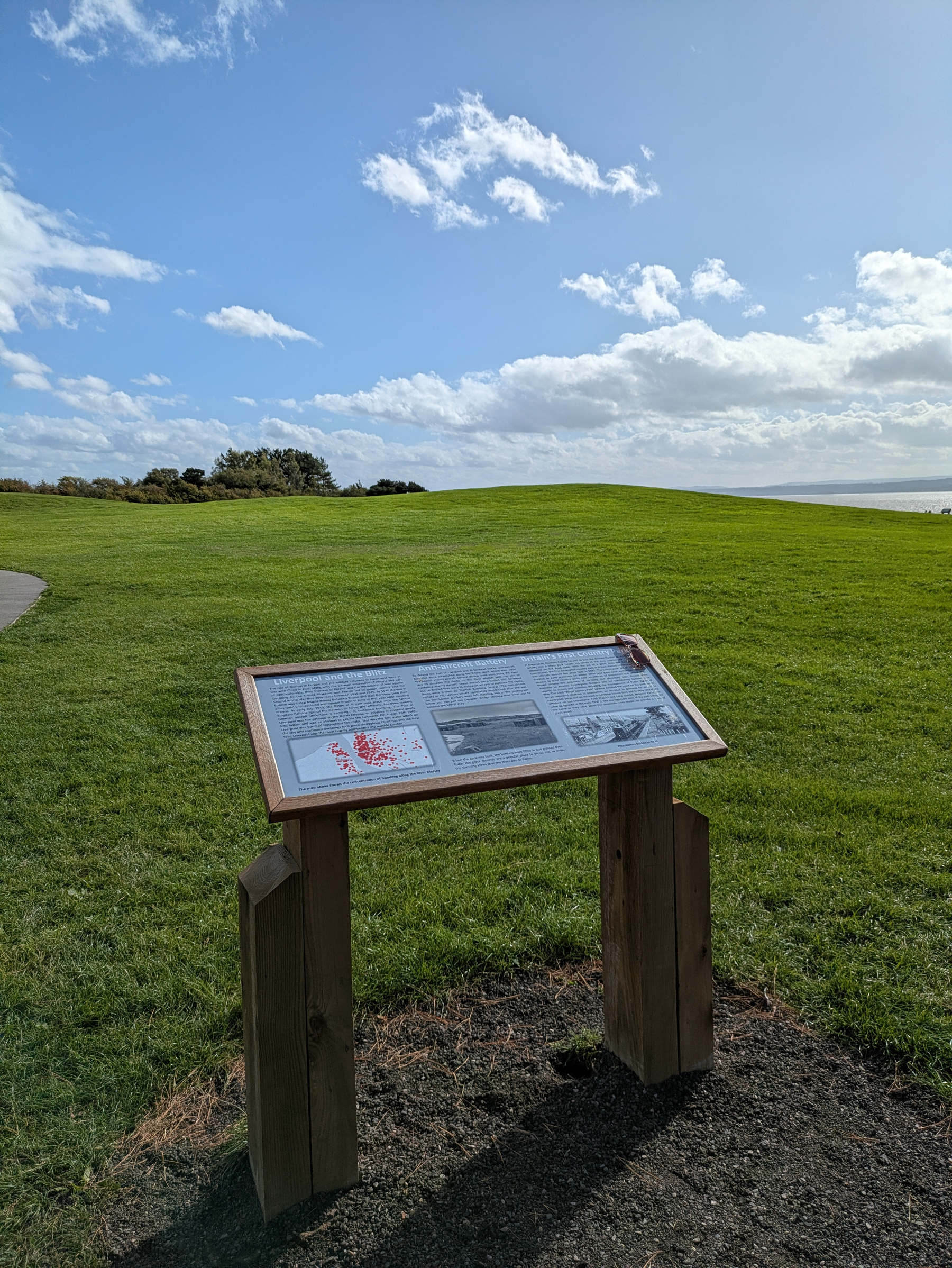 An informational sign is displayed on a wooden stand in a grassy park area under a partly cloudy sky.