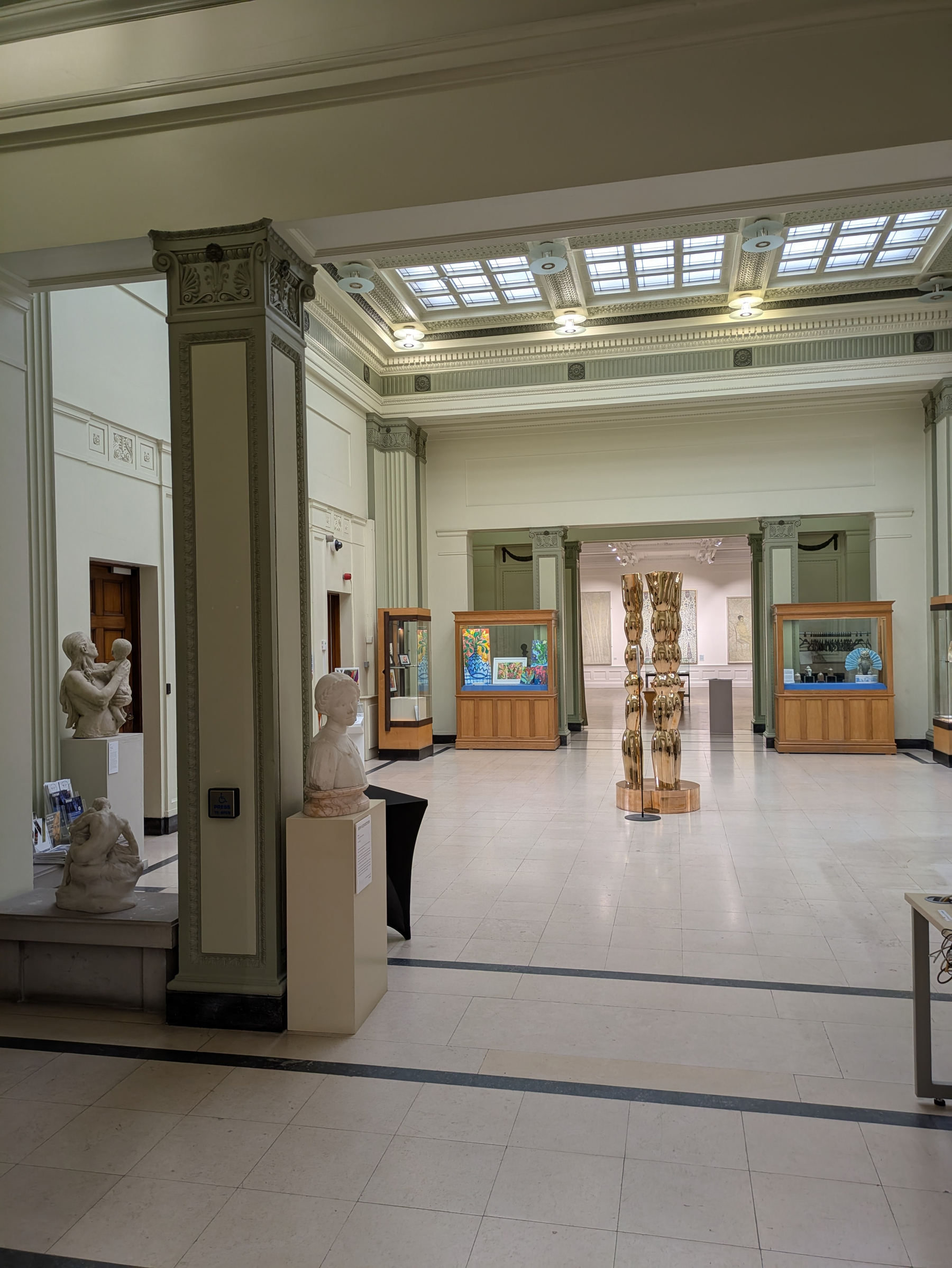 A spacious, well-lit museum gallery features various sculptures, including busts and a tall wooden totem-like piece, along with display cases containing artwork.