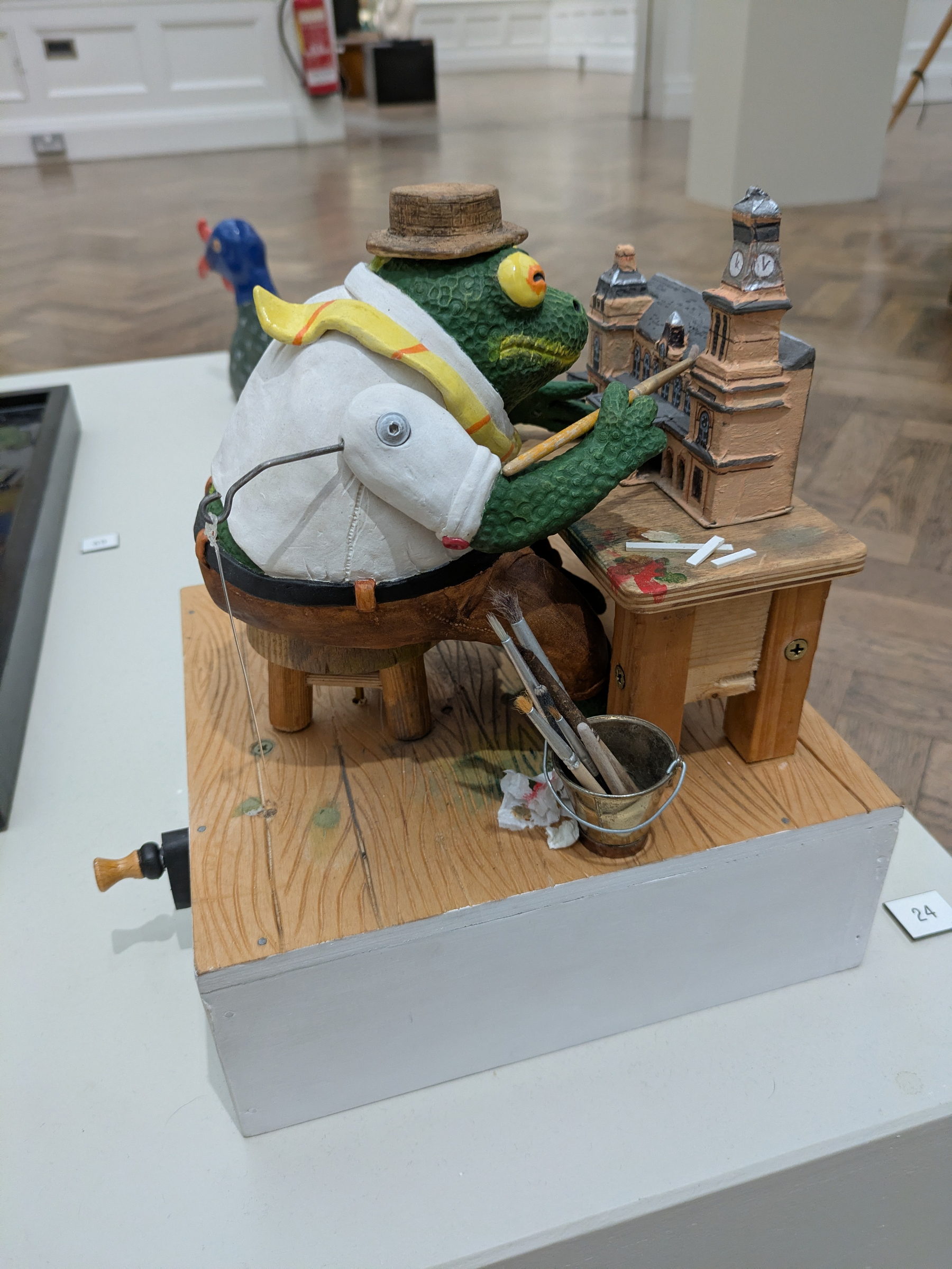 A whimsical frog figure wearing a hat and a shirt sits on a wooden stool, meticulously painting a small architectural model on a desk.
