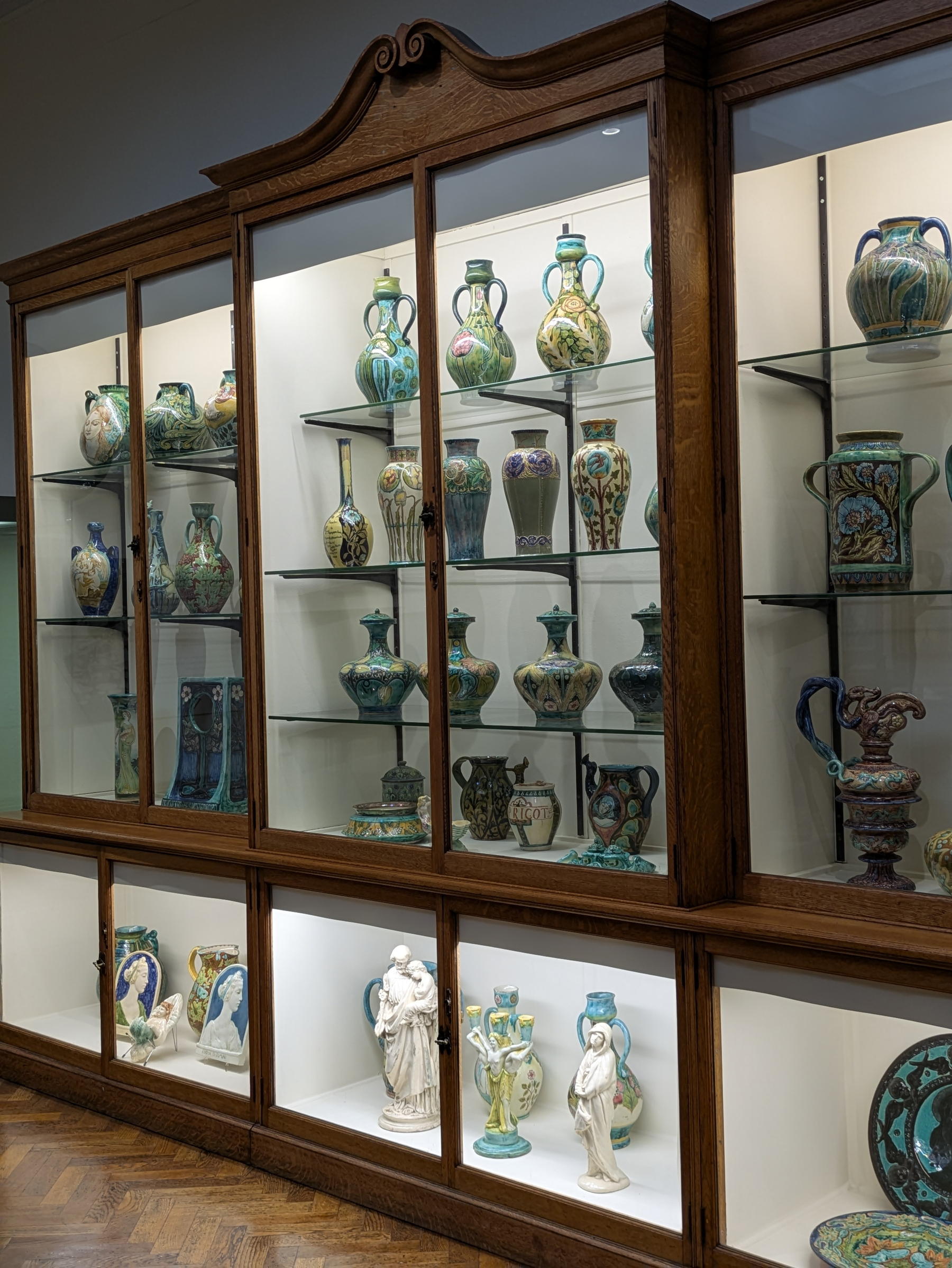 A wooden display cabinet houses a collection of intricately decorated ceramic and porcelain vases, jugs, and figurines, arranged neatly on glass shelves with bright lighting.