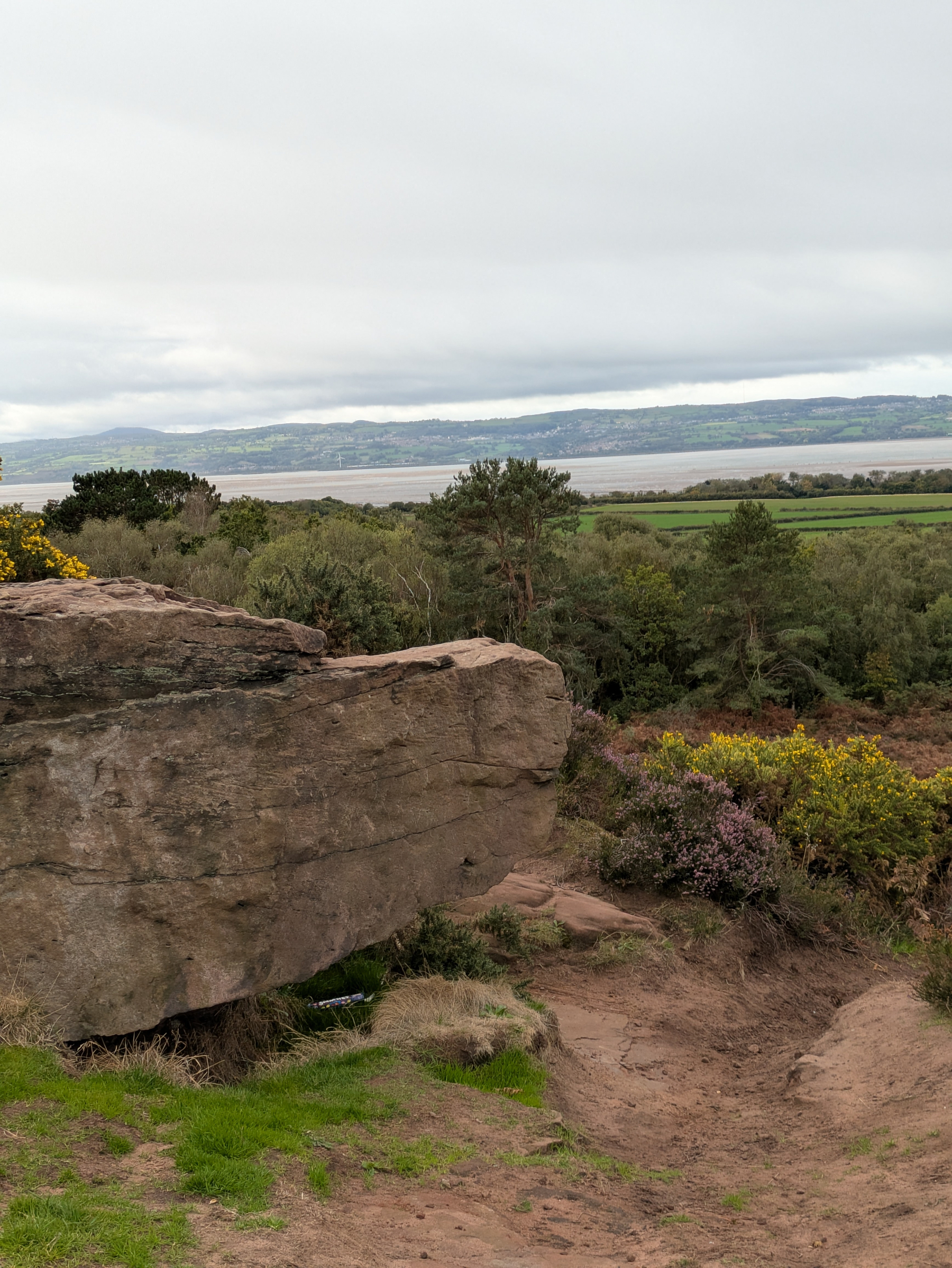 A large rock formation juts out over a dirt path surrounded by greenery, with a view of distant hills and a body of water under a cloudy sky.