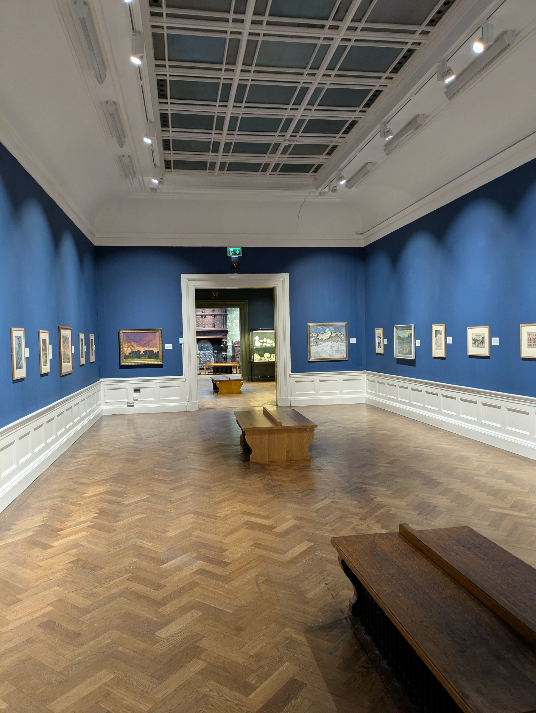 A spacious art gallery room with blue walls displays various framed paintings and drawings, with wooden benches placed in the centre and foreground.