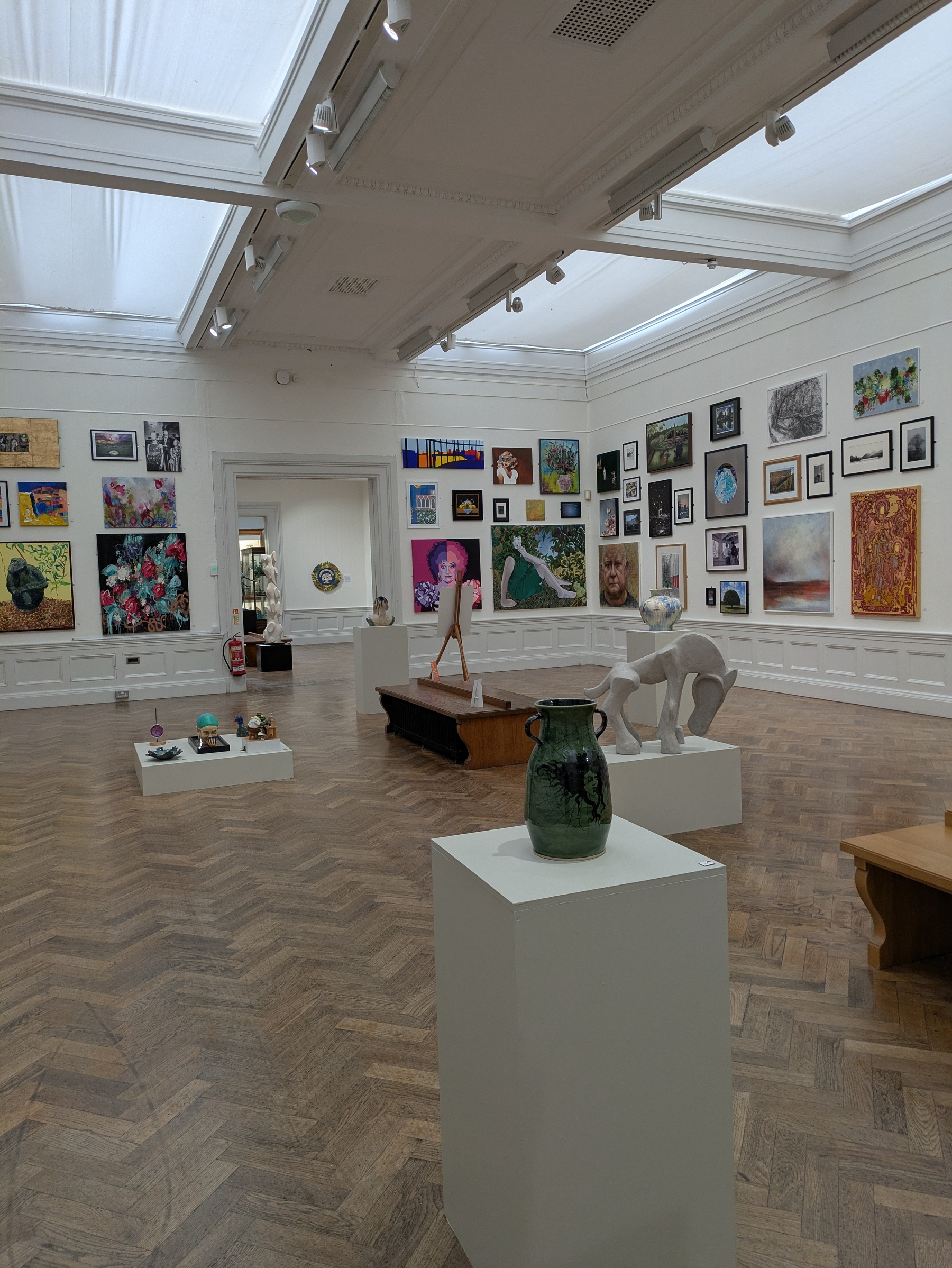 A spacious art gallery displays a variety of paintings on its walls and sculptures on pedestals across a polished wooden floor.