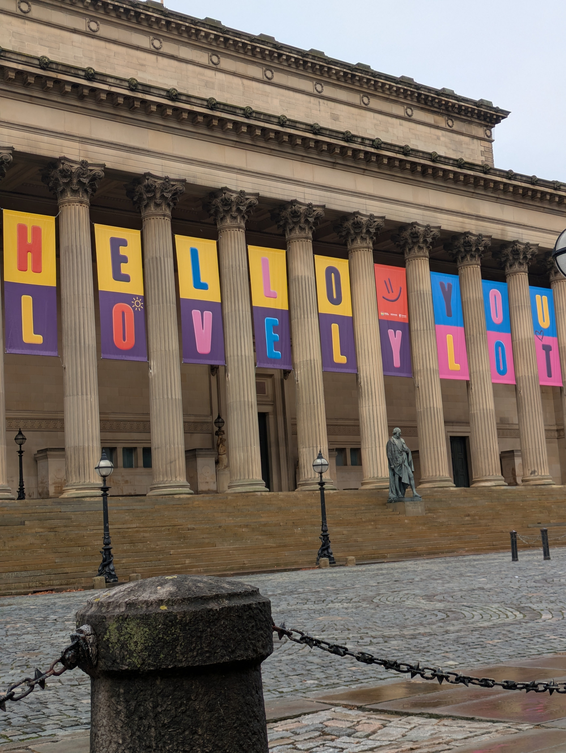 A series of colorful banners spelling out HELLO YOU LOVELY LOT hang between the columns of a grand classical building.