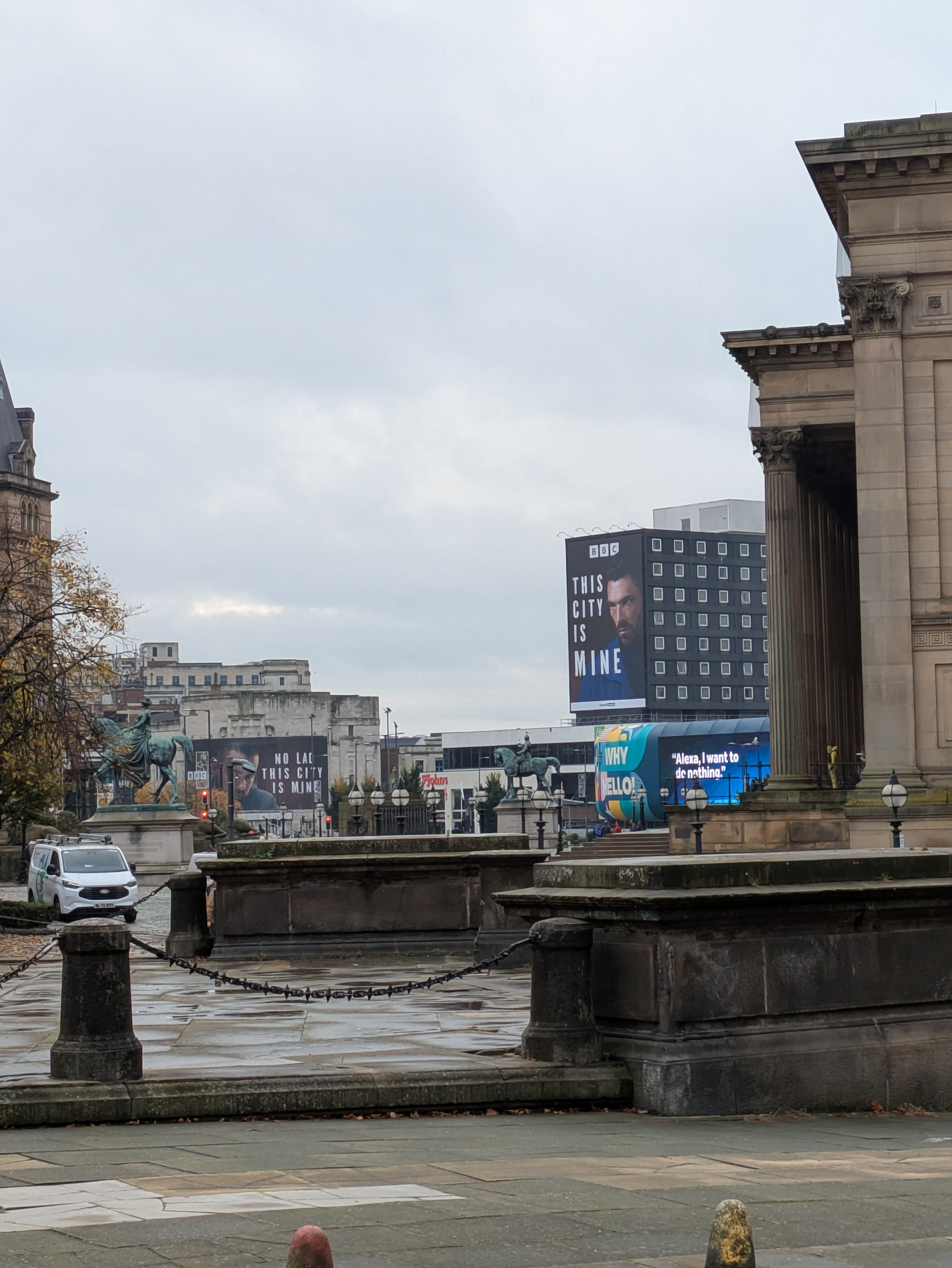 A city square with historic statues and buildings features large digital billboards displaying advertisements and messages.