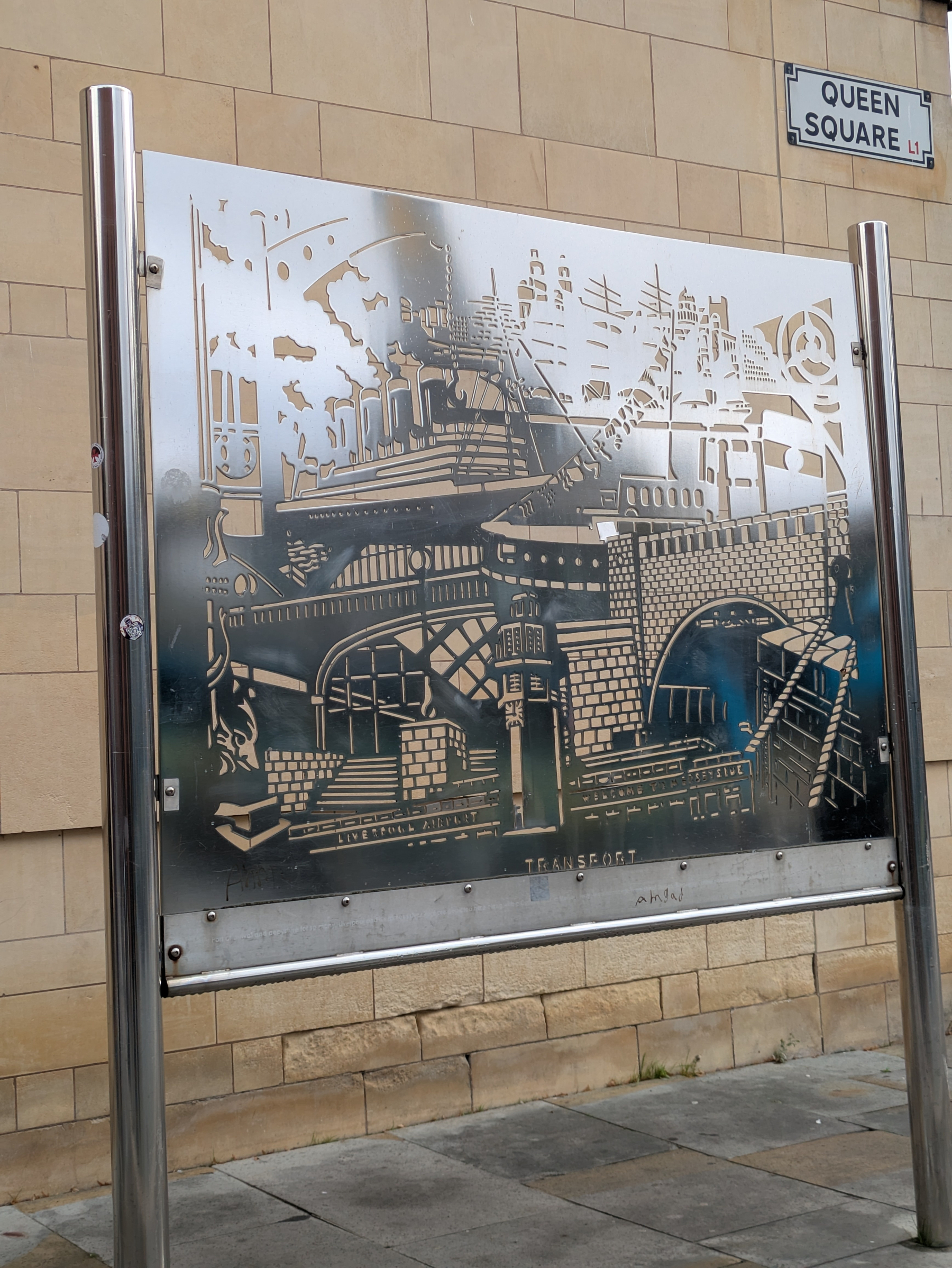 A metallic art panel featuring detailed cut-out designs of various transportation elements, mounted on poles in front of a beige brick wall near a street sign for Queen Square.