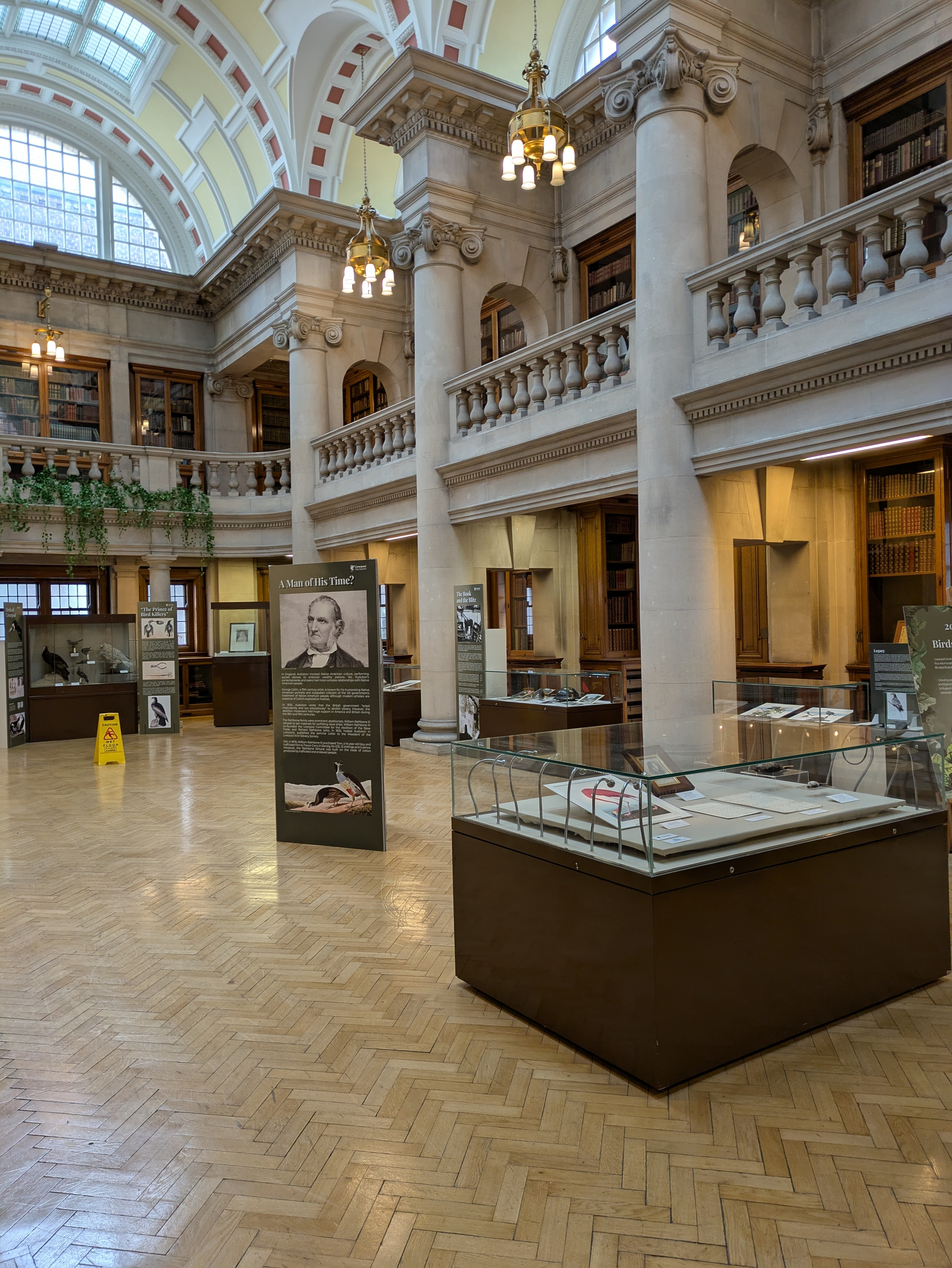 A spacious, historic library or museum interior features tall columns, ornate balconies, display cases with exhibits, and a large skylight ceiling.