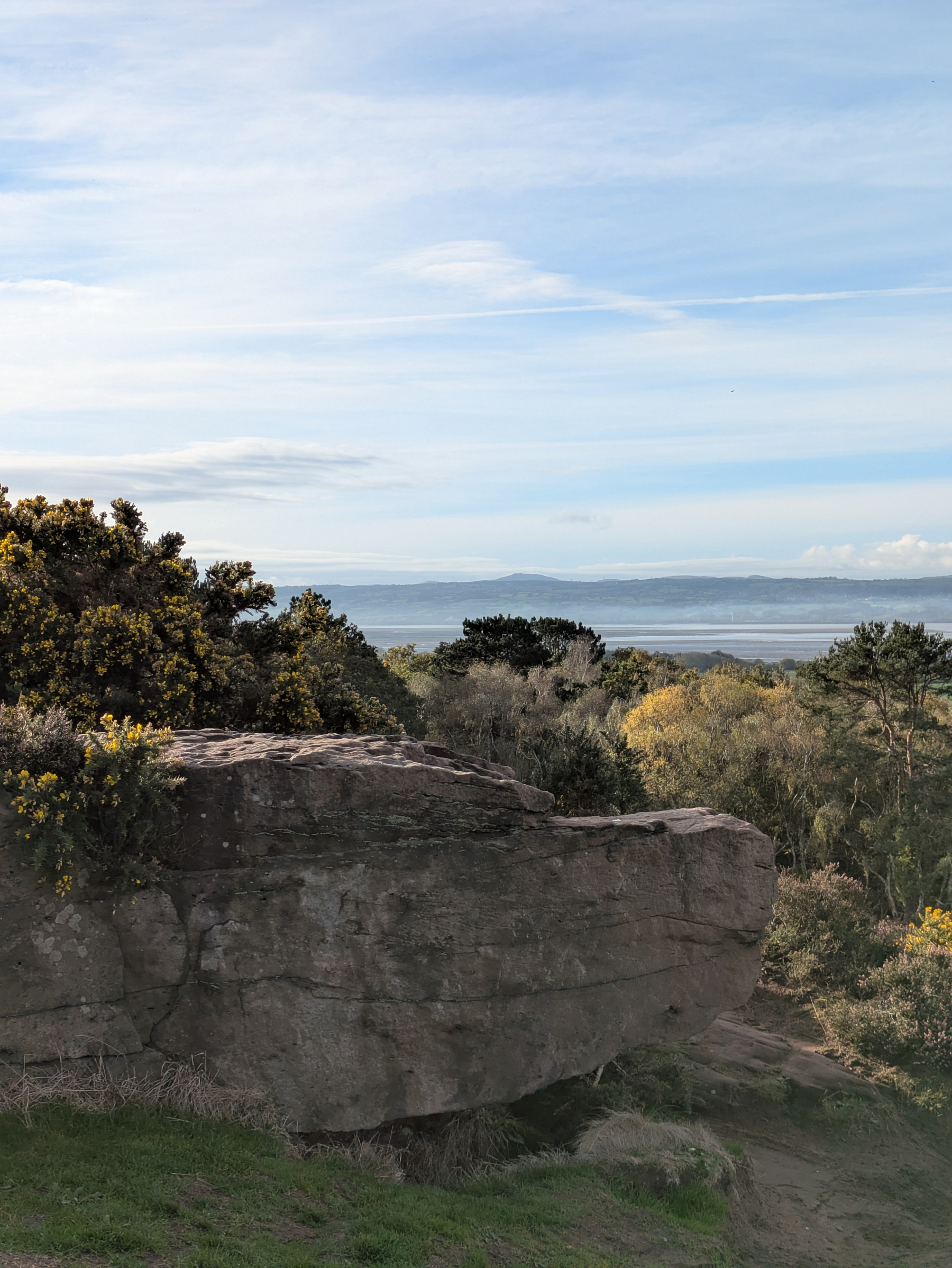 A large rock formation is surrounded by dense greenery with a view of distant hills and a body of water under a partly cloudy sky.