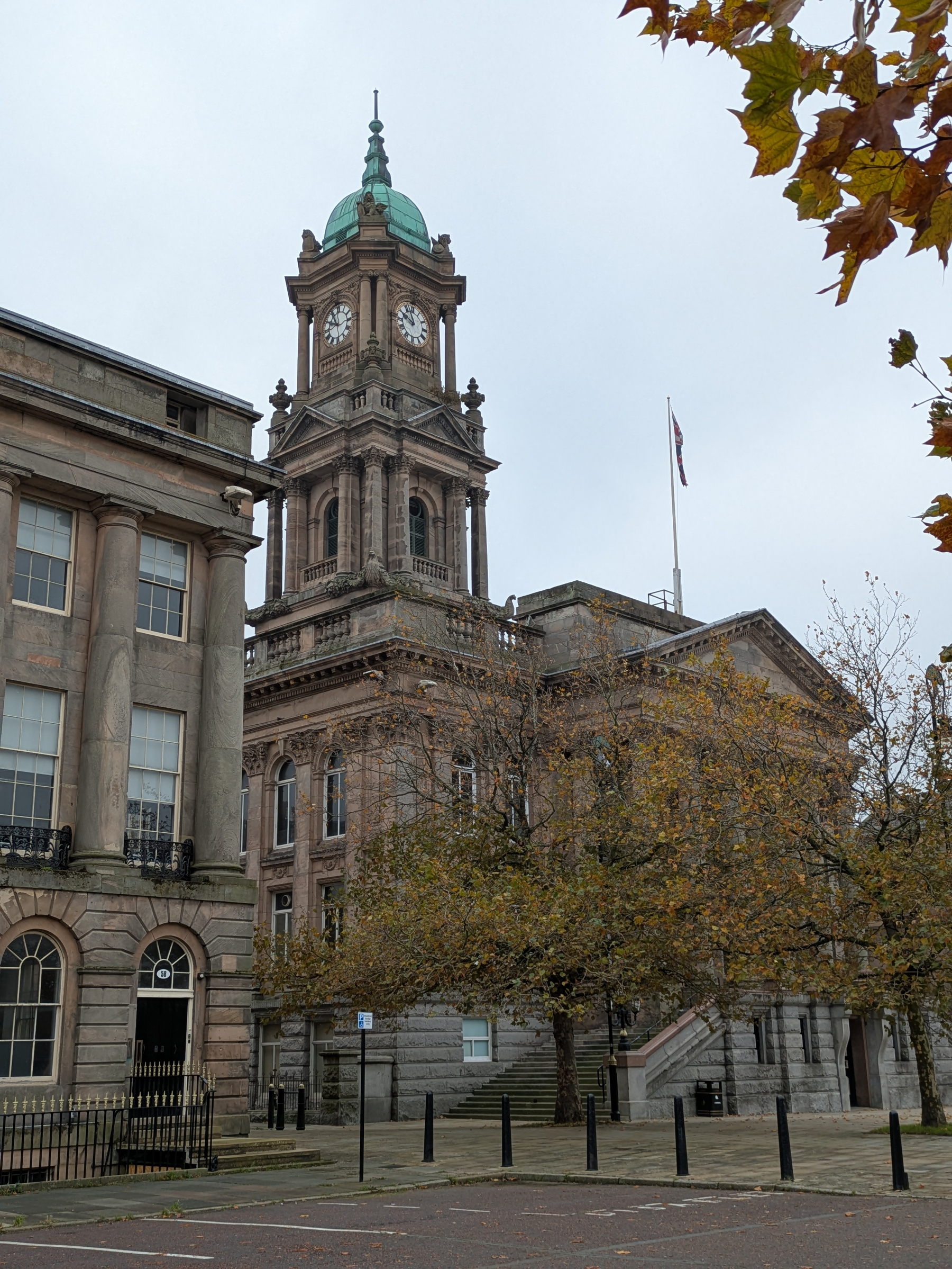 A historic building with a clock tower and a green dome is surrounded by trees with autumn leaves on a cloudy day.