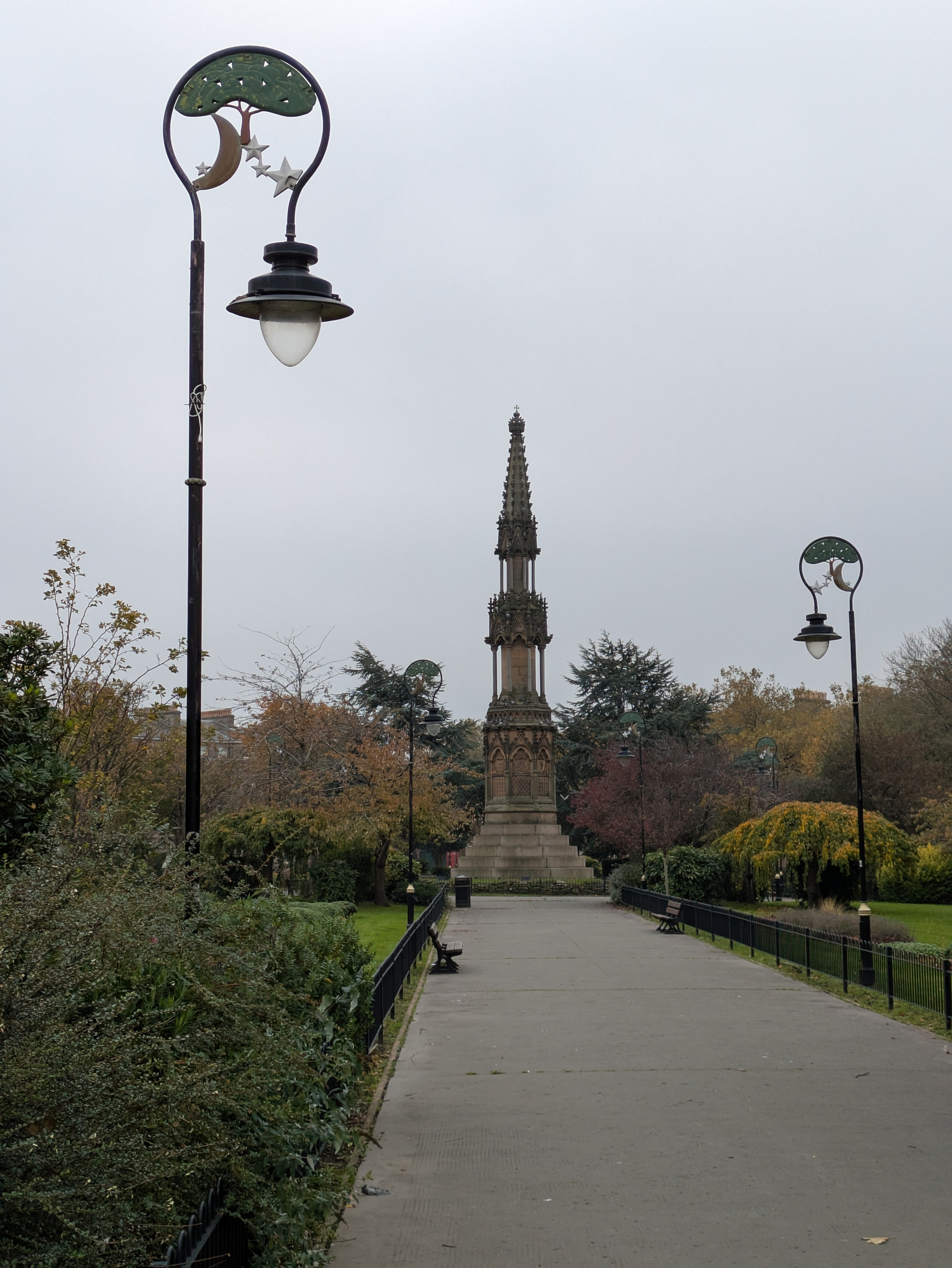 A paved pathway lined with decorative lampposts featuring crescent moon and star designs leads to a tall, ornate stone monument surrounded by trees and greenery.