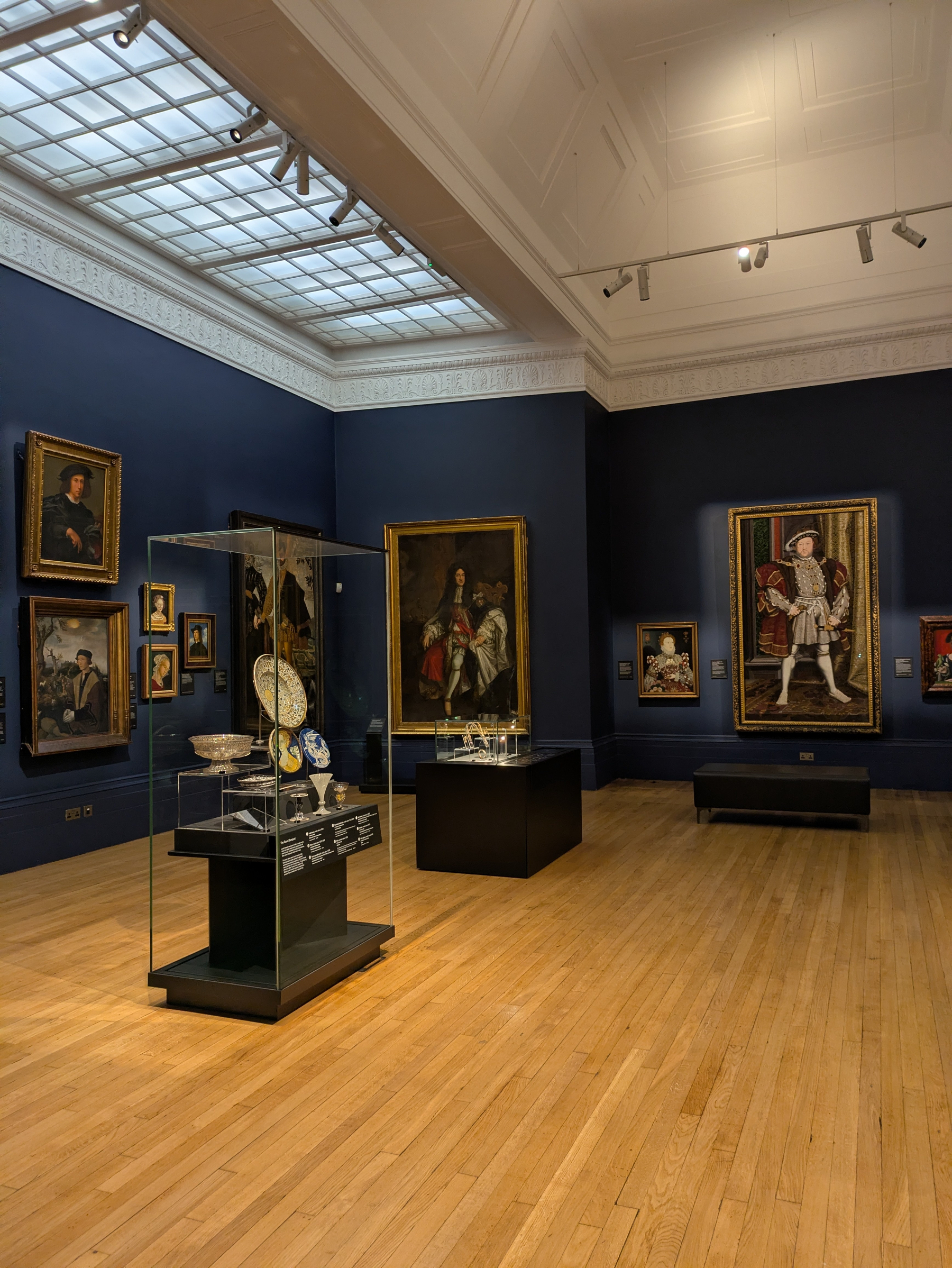 A gallery room features paintings on dark blue walls, with glass display cases holding various artifacts on wooden flooring.