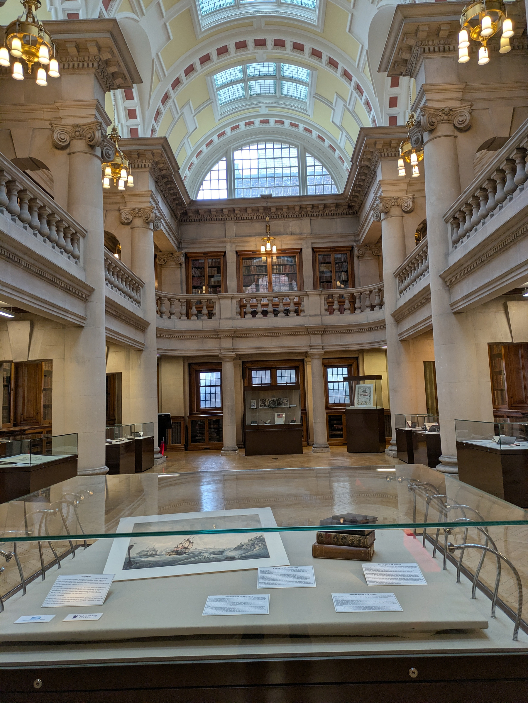 A grand, ornate library interior features tall columns, arched windows, and display cases with books and artifacts.