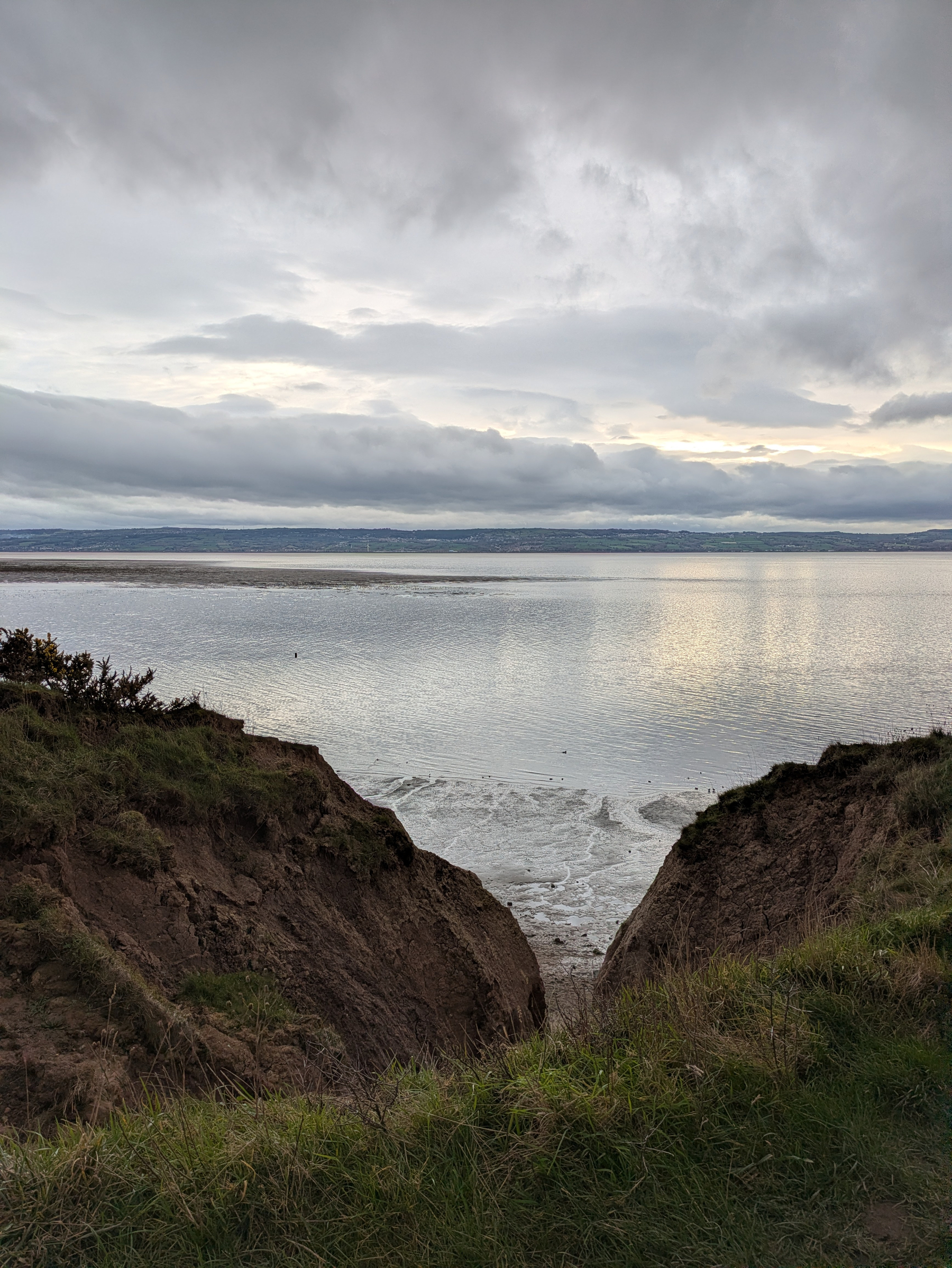 A dramatic coastal view features a grassy cliff opening onto a calm sea under a cloudy sky.