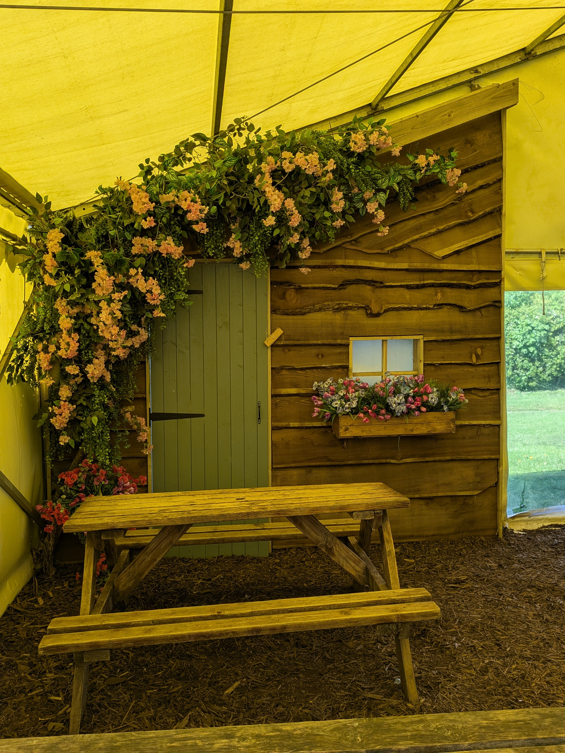 A rustic wooden structure adorned with flowers houses a green door and window, with a wooden picnic table in front under a yellow tent.
