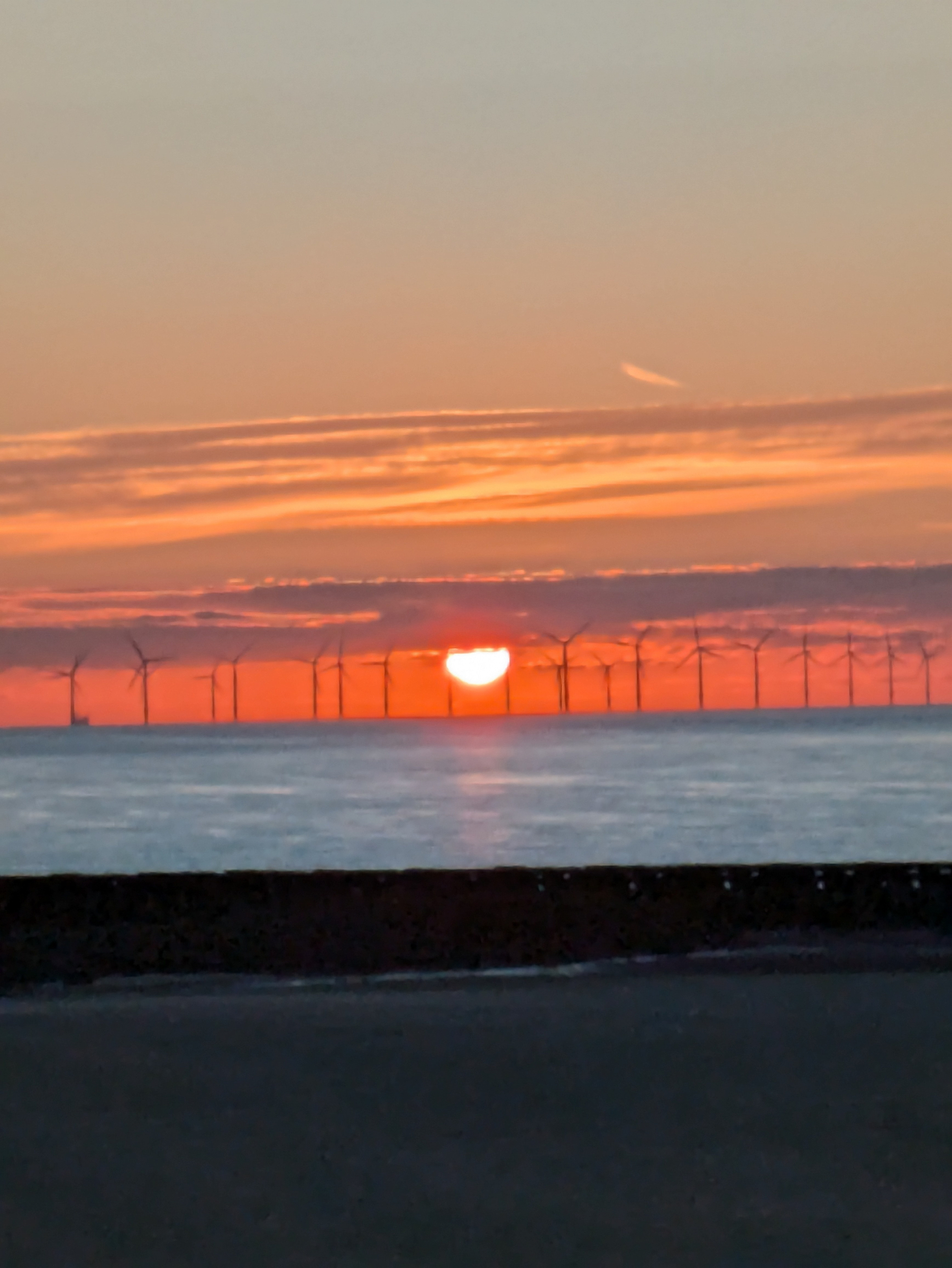 A row of offshore wind turbines stands silhouetted against a vibrant sunset over the ocean.