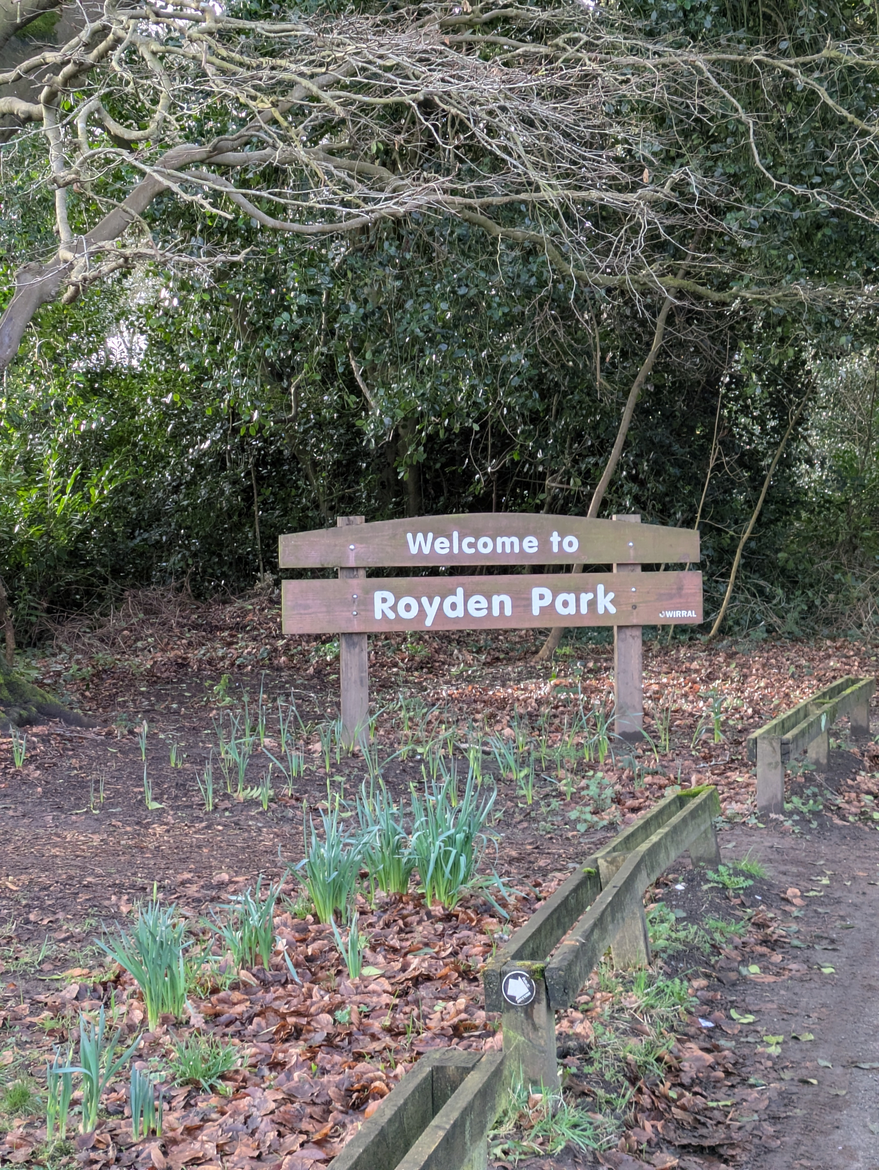 A wooden sign saying Welcome to Royden Park is surrounded by trees, fallen leaves, and emerging plants.