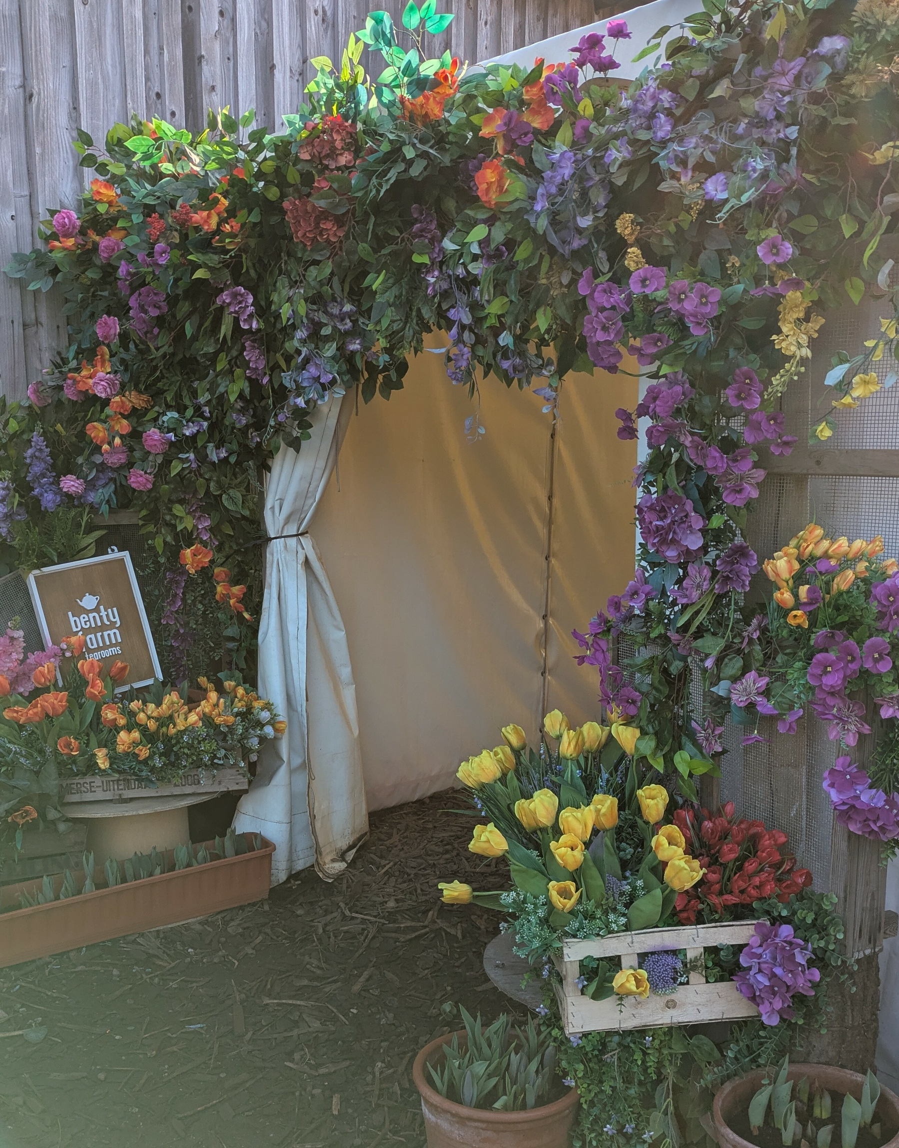 A decorative floral arch adorned with vibrant flowers leads to a cozy tent area, surrounded by colorful flower arrangements.