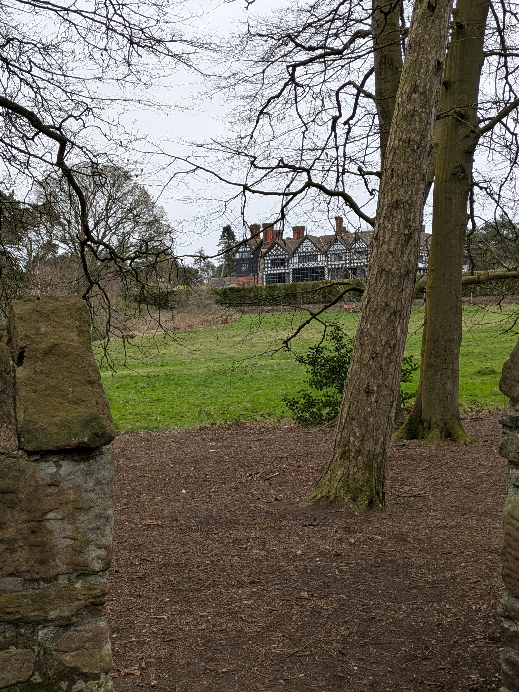 A view of a large, traditional house with a distinctive black-and-white timber design is visible beyond a wooded area and grassy field.