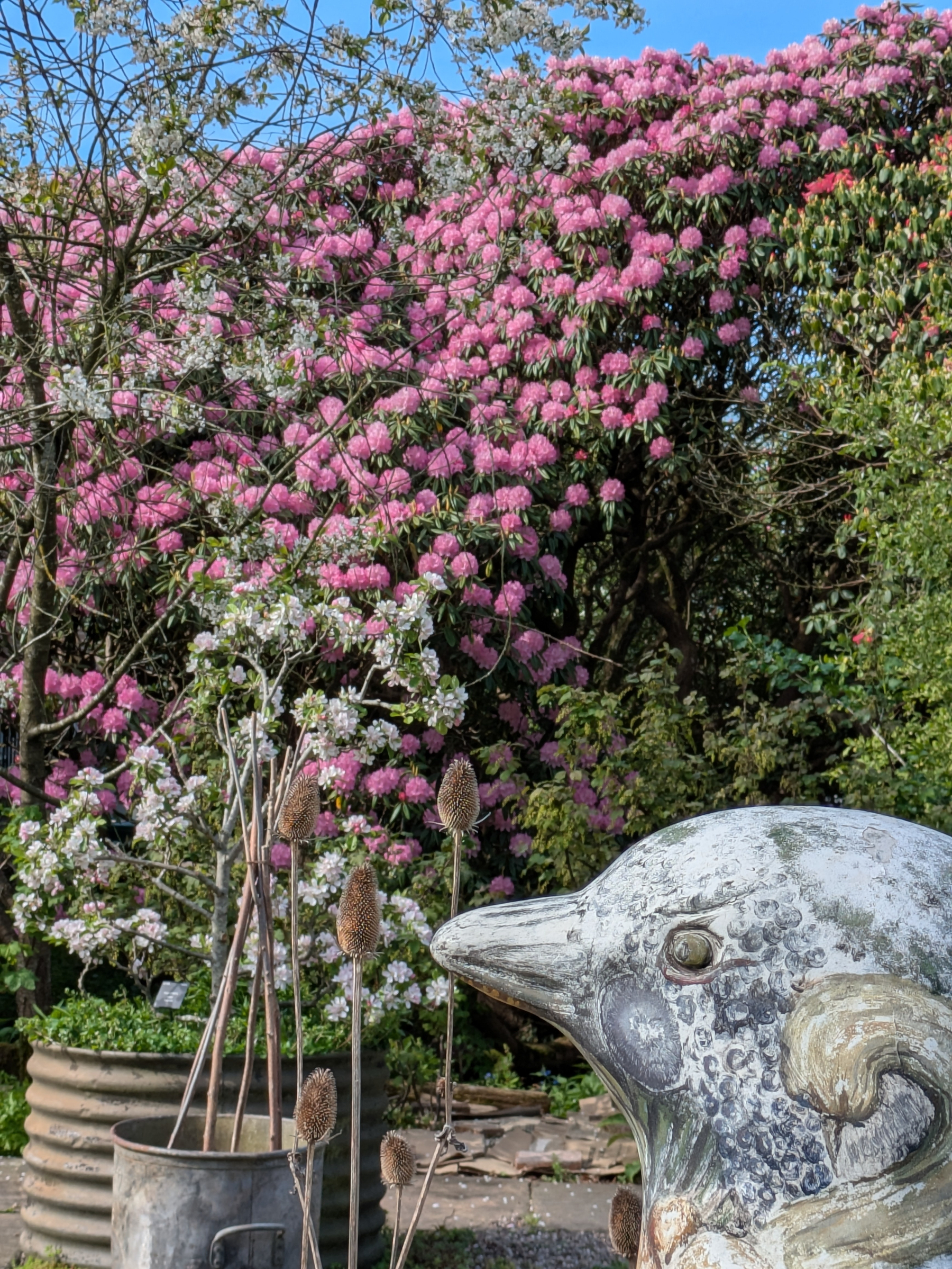 A garden scene features a bird sculpture in the foreground, with blooming pink flowers and trees in the background.