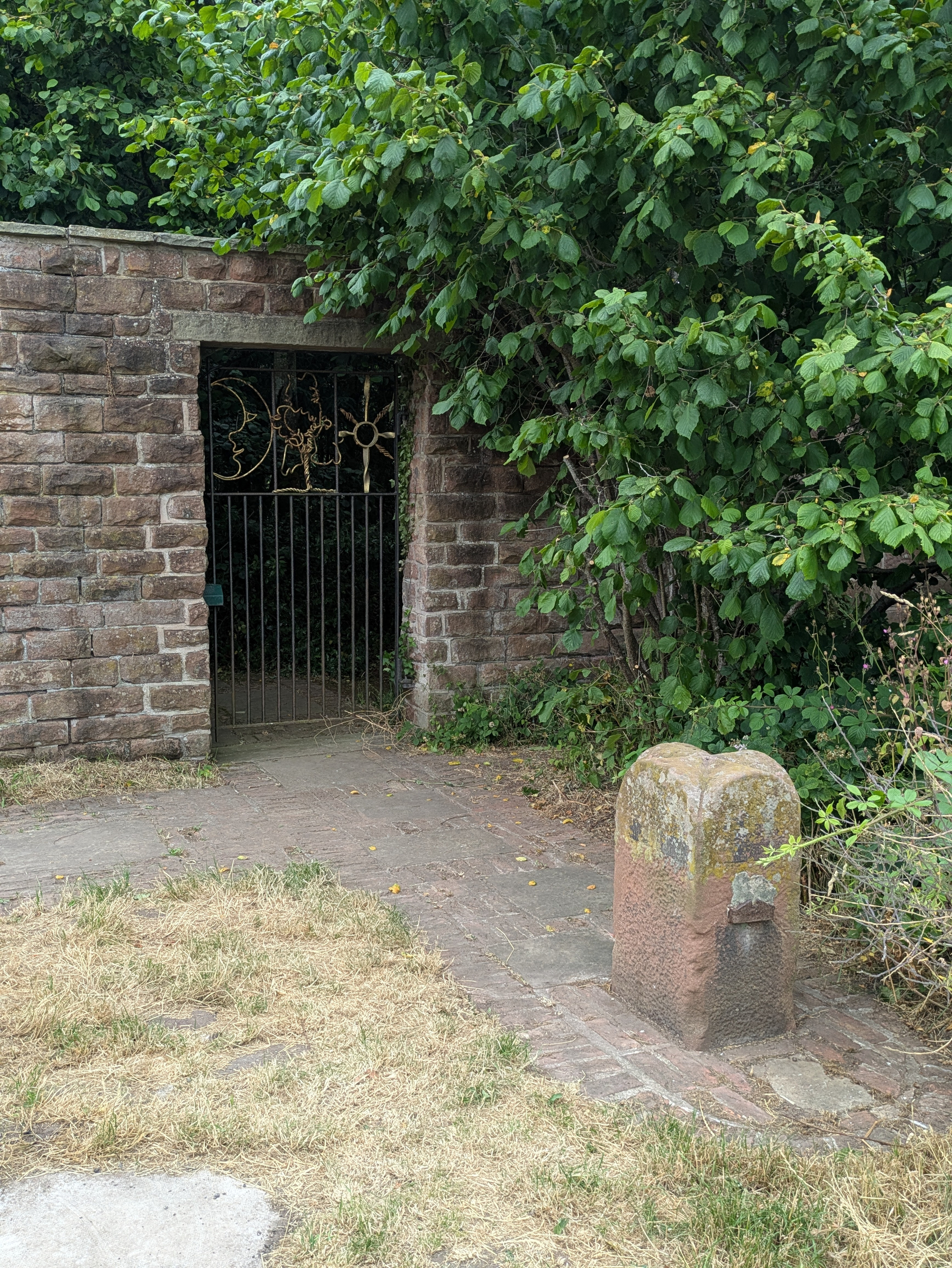 A stone pathway leads to an iron gate adorned with sun and moon designs, set in a brick wall partially covered by lush green foliage.