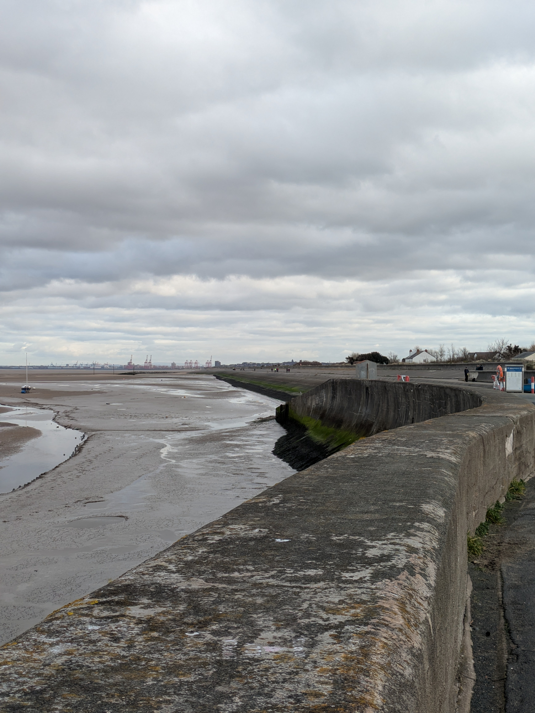 A concrete seawall overlooks a cloudy coastline with a sandy beach and distant industrial structures.
