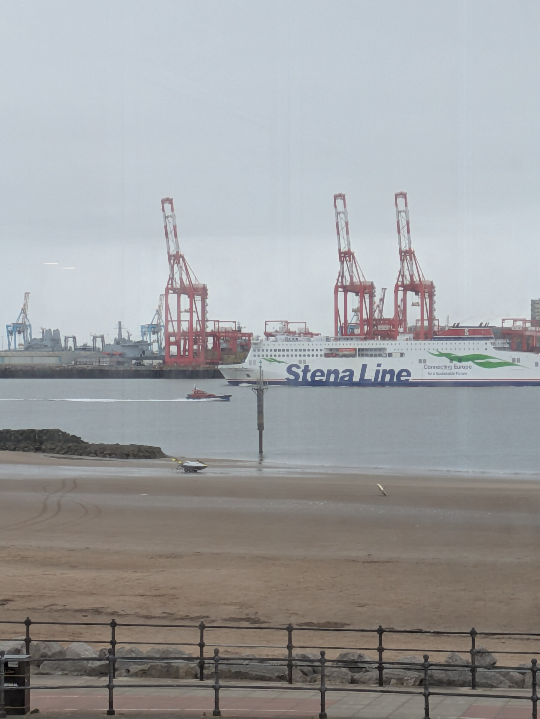 A large passenger ferry labeled Stena Line is passing near a wharf with cranes visible in the background and a sandy beach in the foreground.