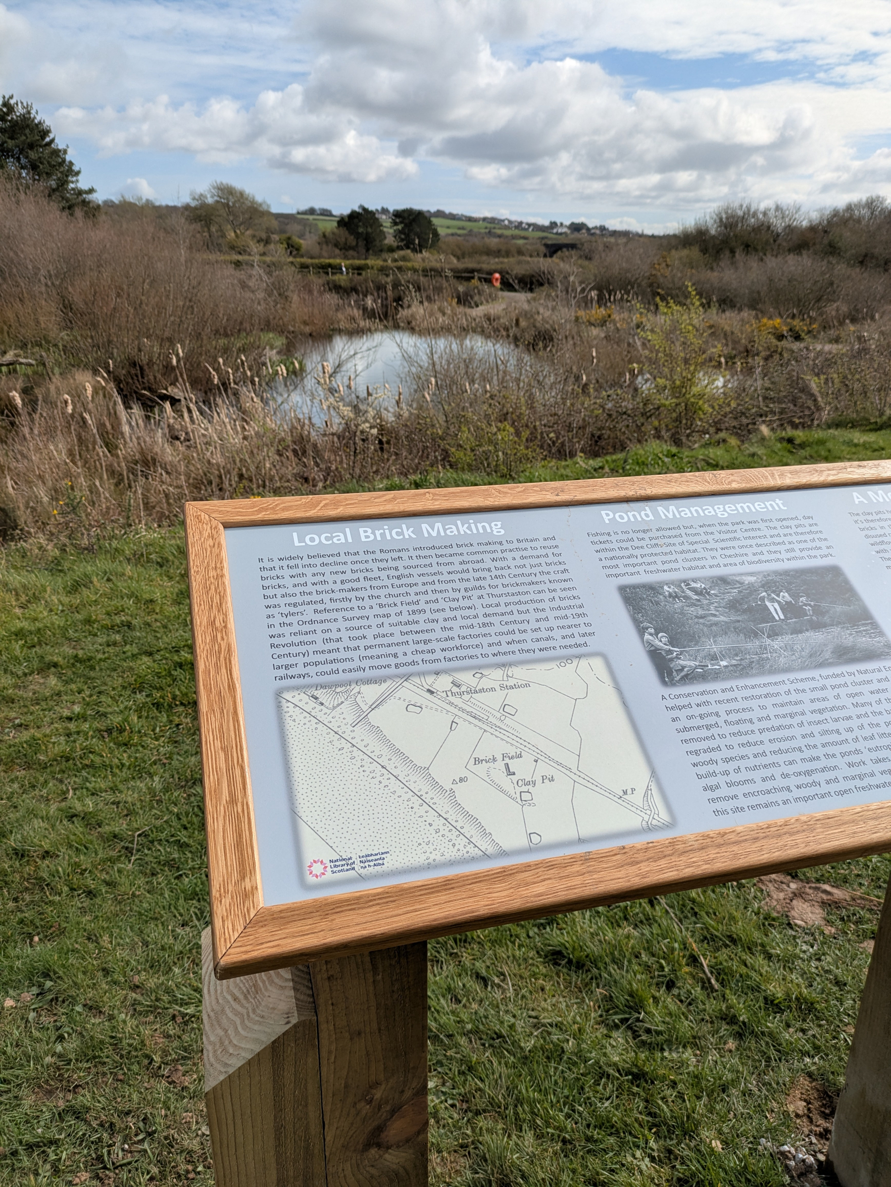 A wooden information board about local brick making and pond management stands in front of a scenic landscape with a pond and grassy area.