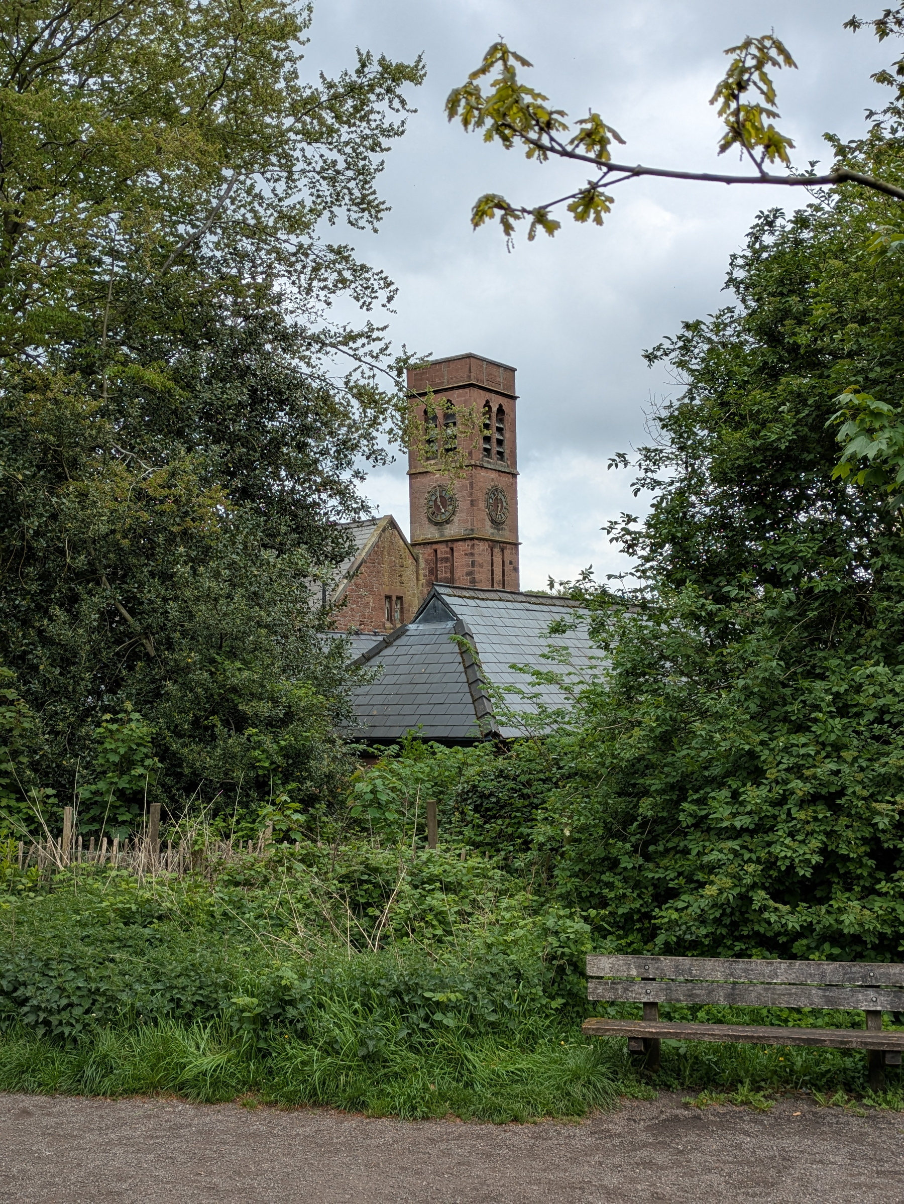 A brick tower rises above lush greenery, with a pathway and bench in the foreground.