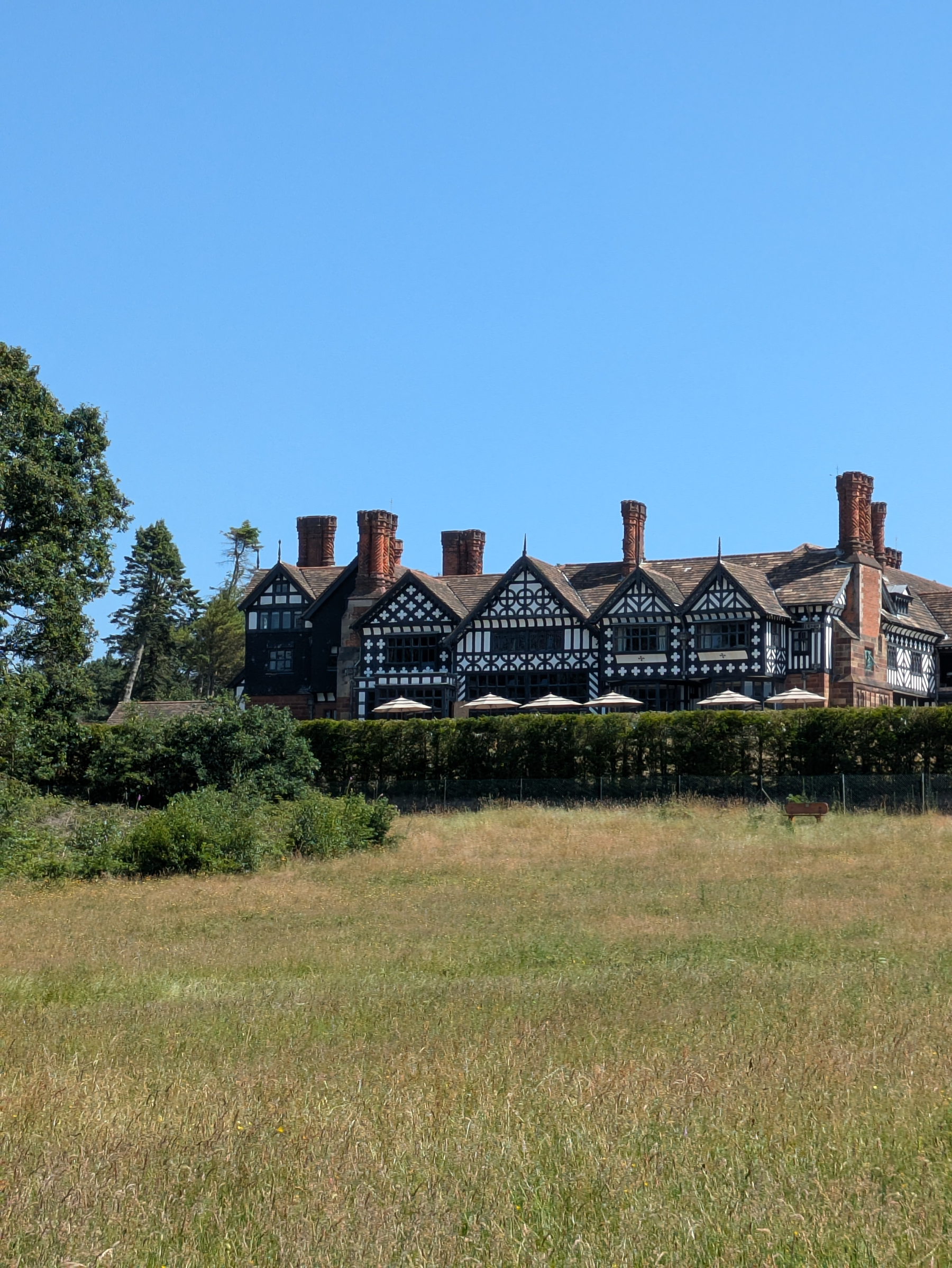 A large, Tudor-style manor house with black and white timber framing and multiple chimneys is set behind a green field under a clear blue sky.